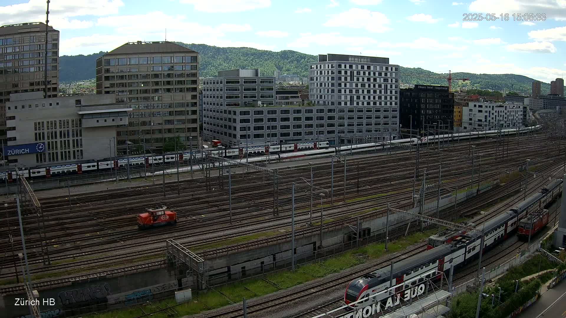 A sunny day reveals a sprawling railway yard with multiple trains, interspersed with modern buildings and a backdrop of green hills.
