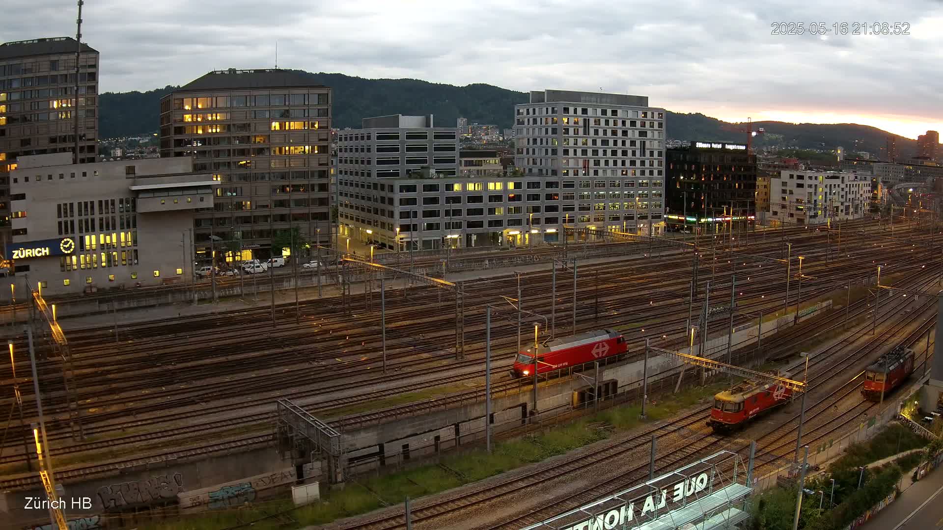 A high-angle, evening view of a large train yard with several trains, surrounded by city buildings under a cloudy sky.