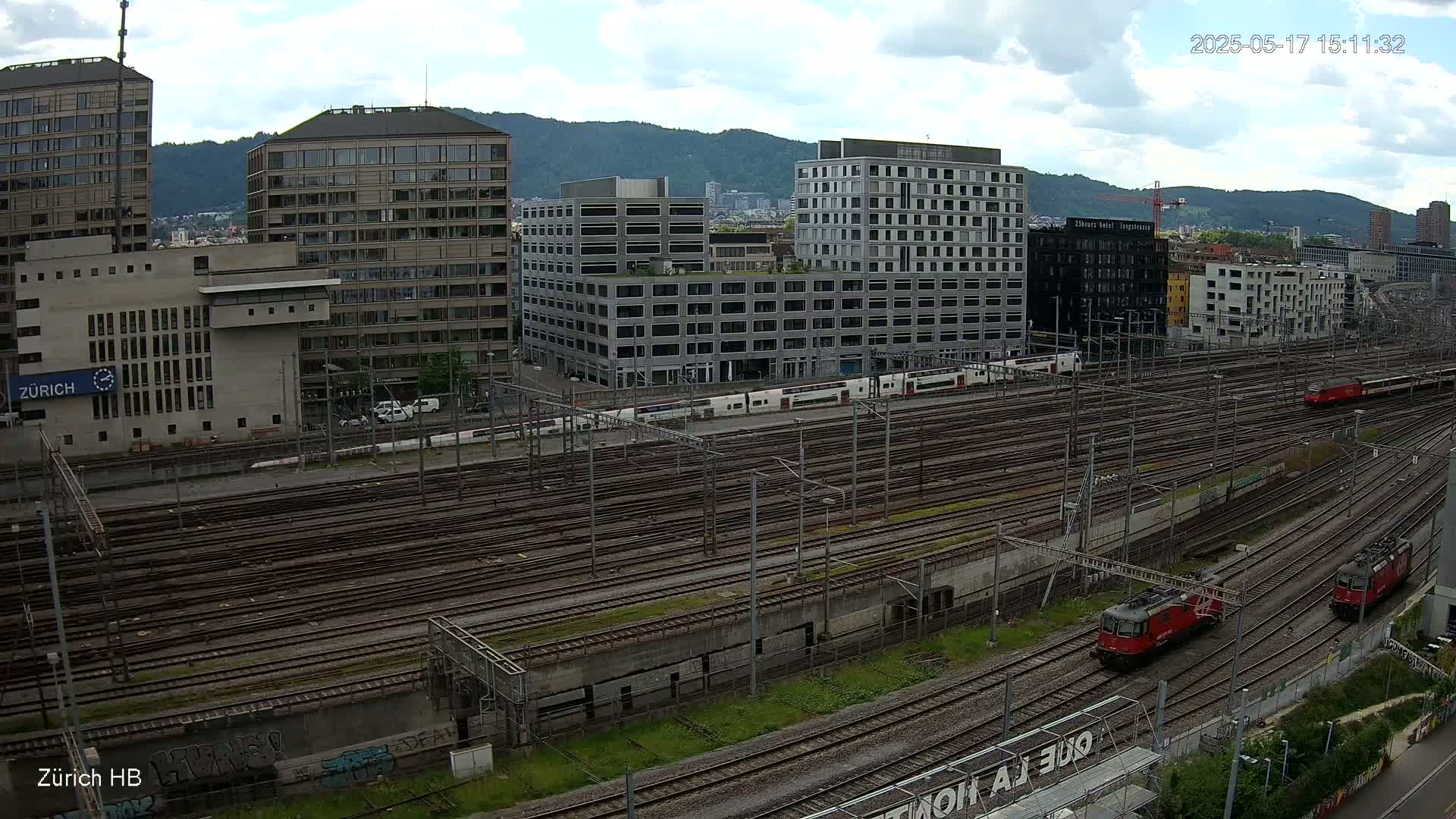 A sprawling railway yard with numerous tracks and several trains, surrounded by modern buildings under a partly cloudy sky.