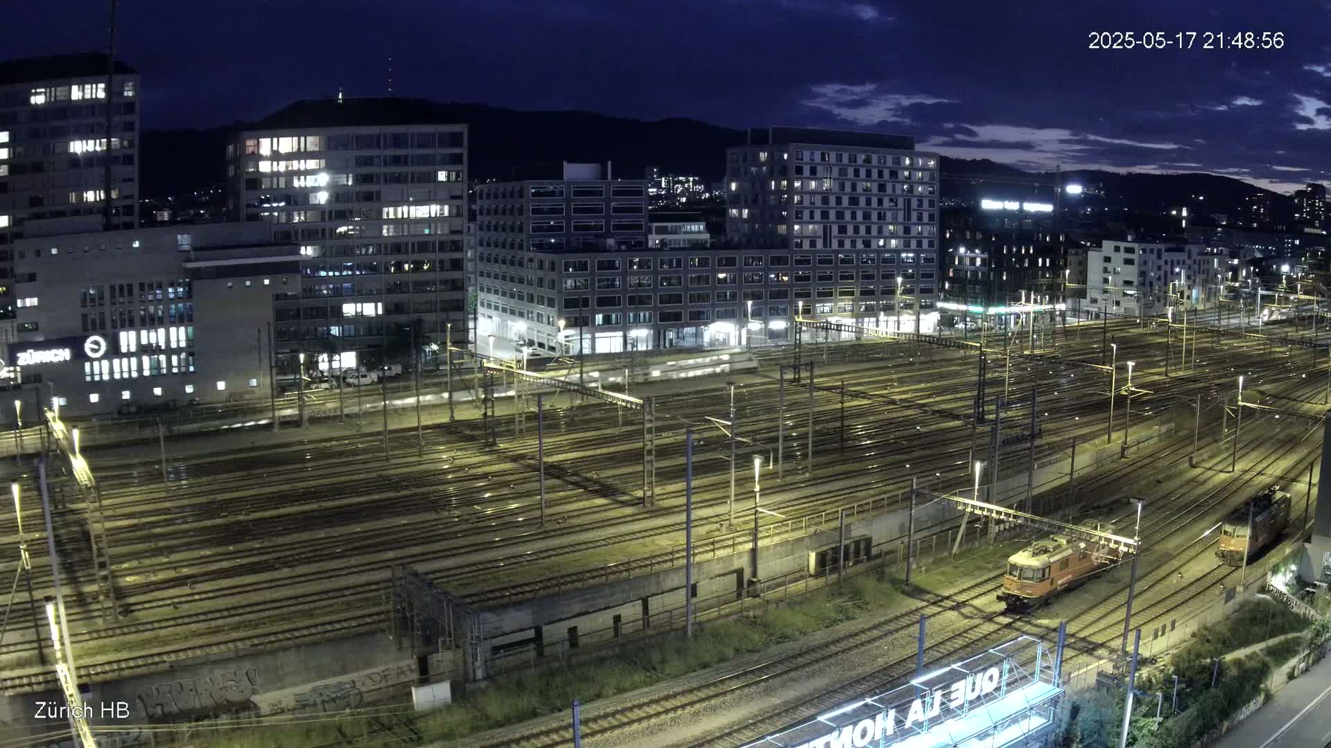 A nighttime aerial view of a large railway yard surrounded by illuminated buildings under a partly cloudy sky.