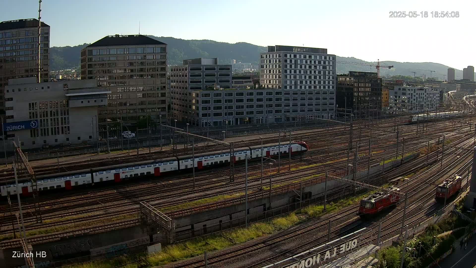 A long passenger train and two locomotives are visible on a sprawling network of train tracks next to several multi-story buildings under a clear, sunny sky.