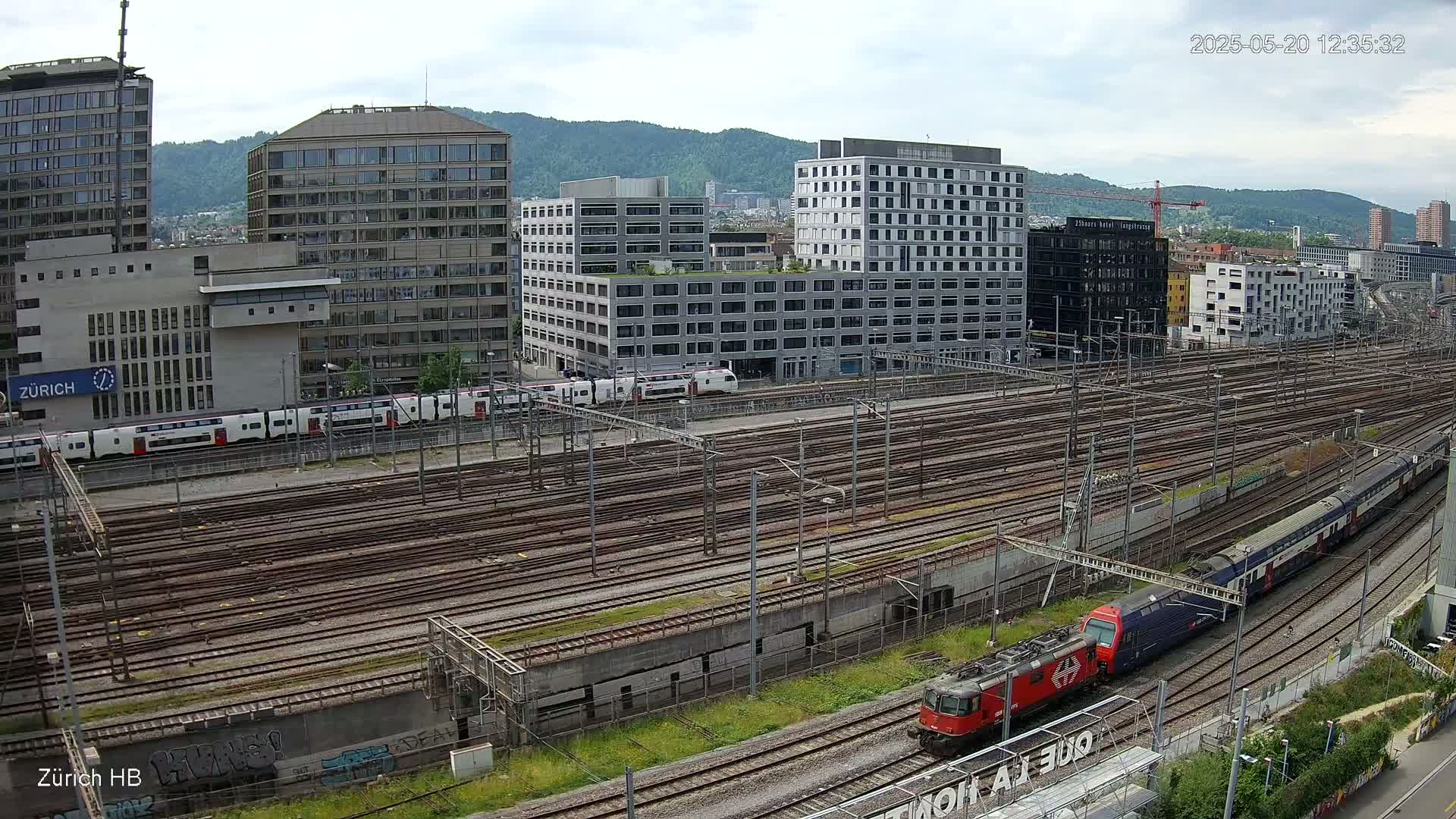 A high-angle view of a railway yard with numerous train tracks, surrounded by modern buildings under a cloudy sky, showing a train passing through.