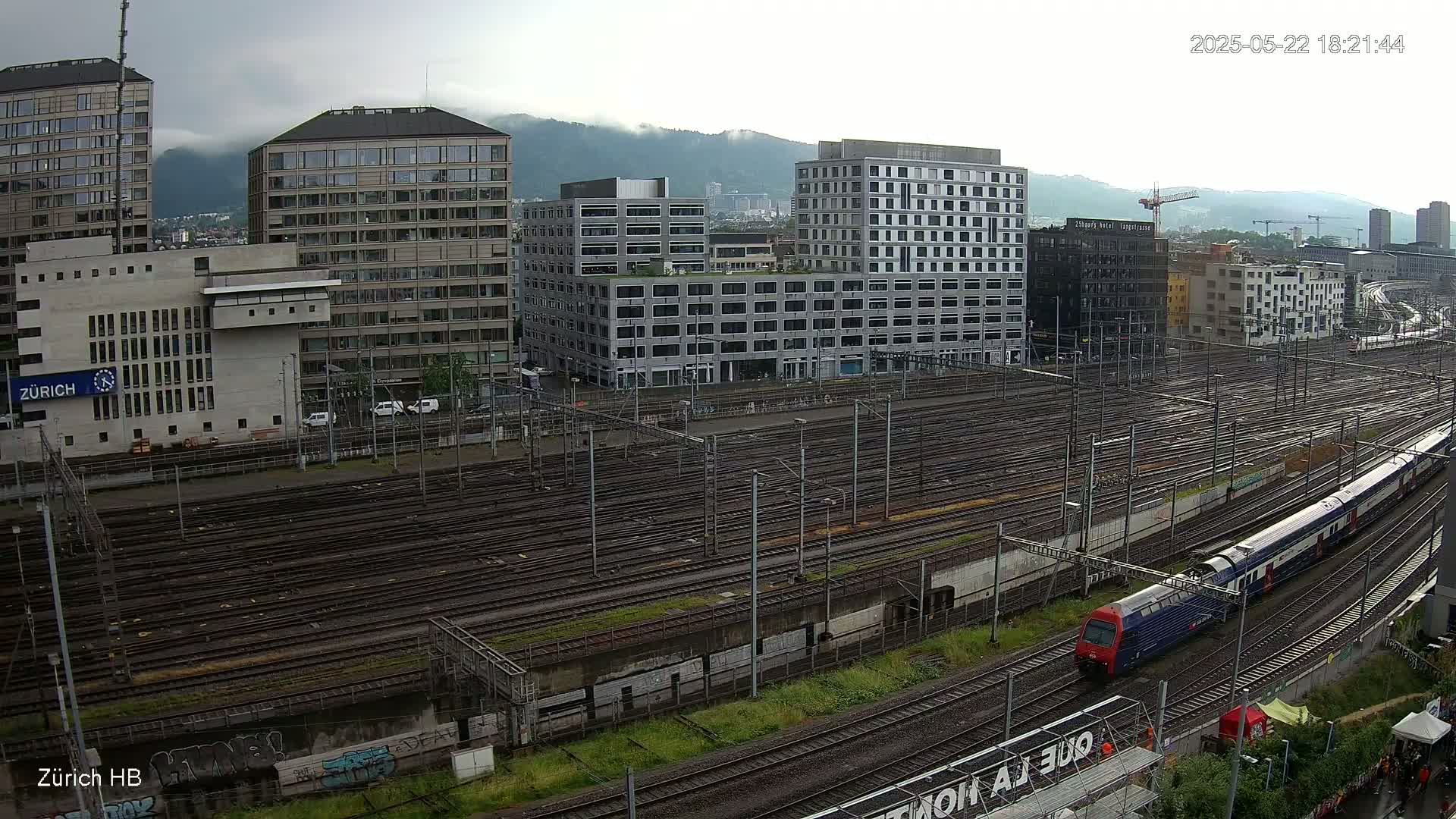 A train passes through a sprawling railway yard next to several modern buildings under an overcast sky.