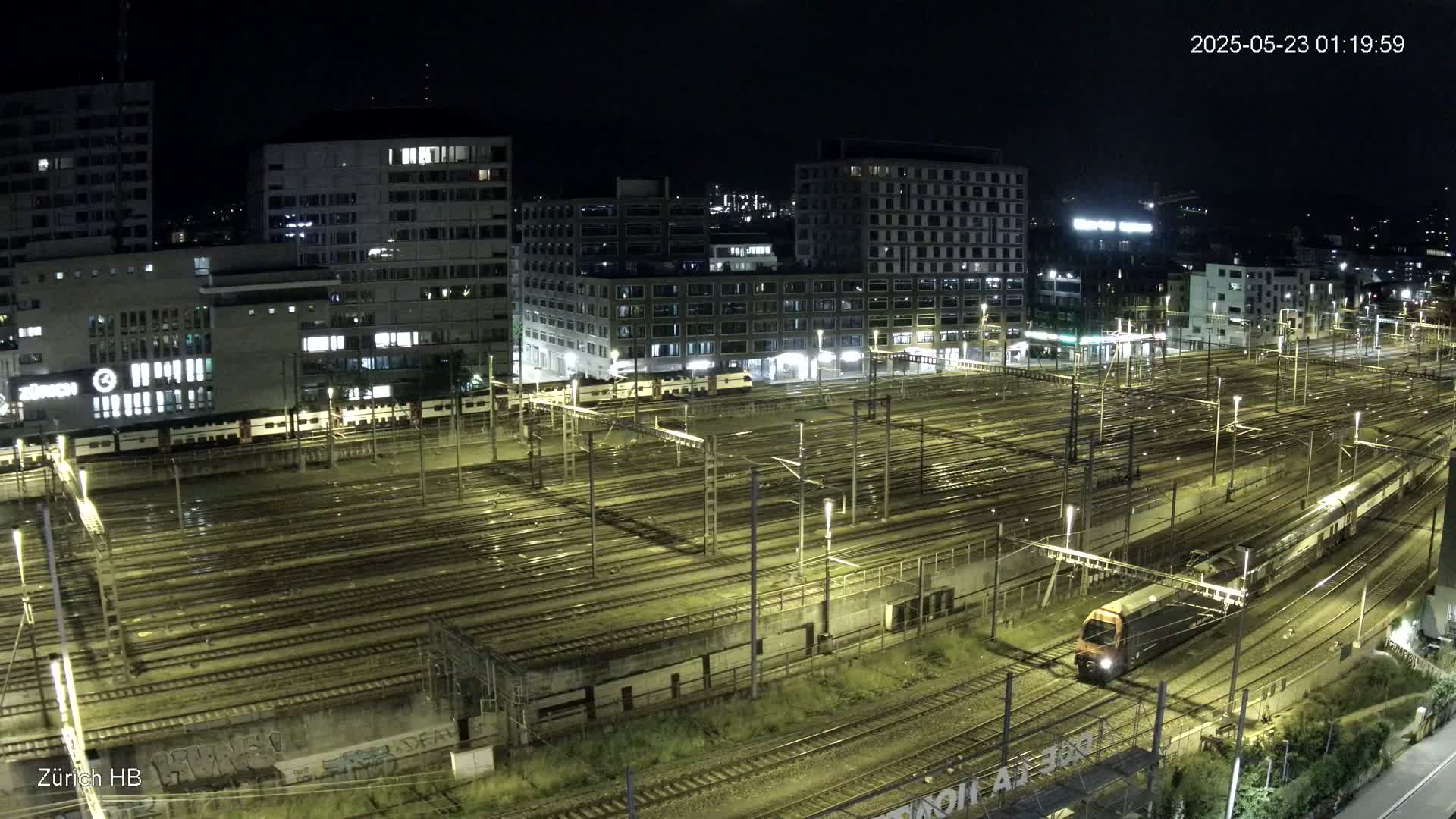 A nighttime aerial view of a train station with numerous train tracks, illuminated buildings, and a train traveling through the station.