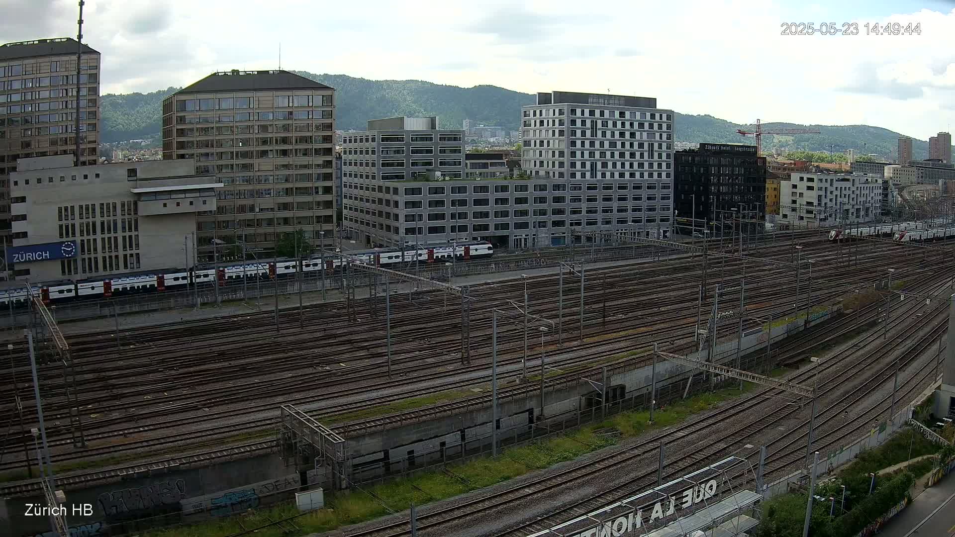 An overcast day reveals a sprawling railway yard filled with numerous train tracks, bordered by several multi-story buildings and a distant mountain range.