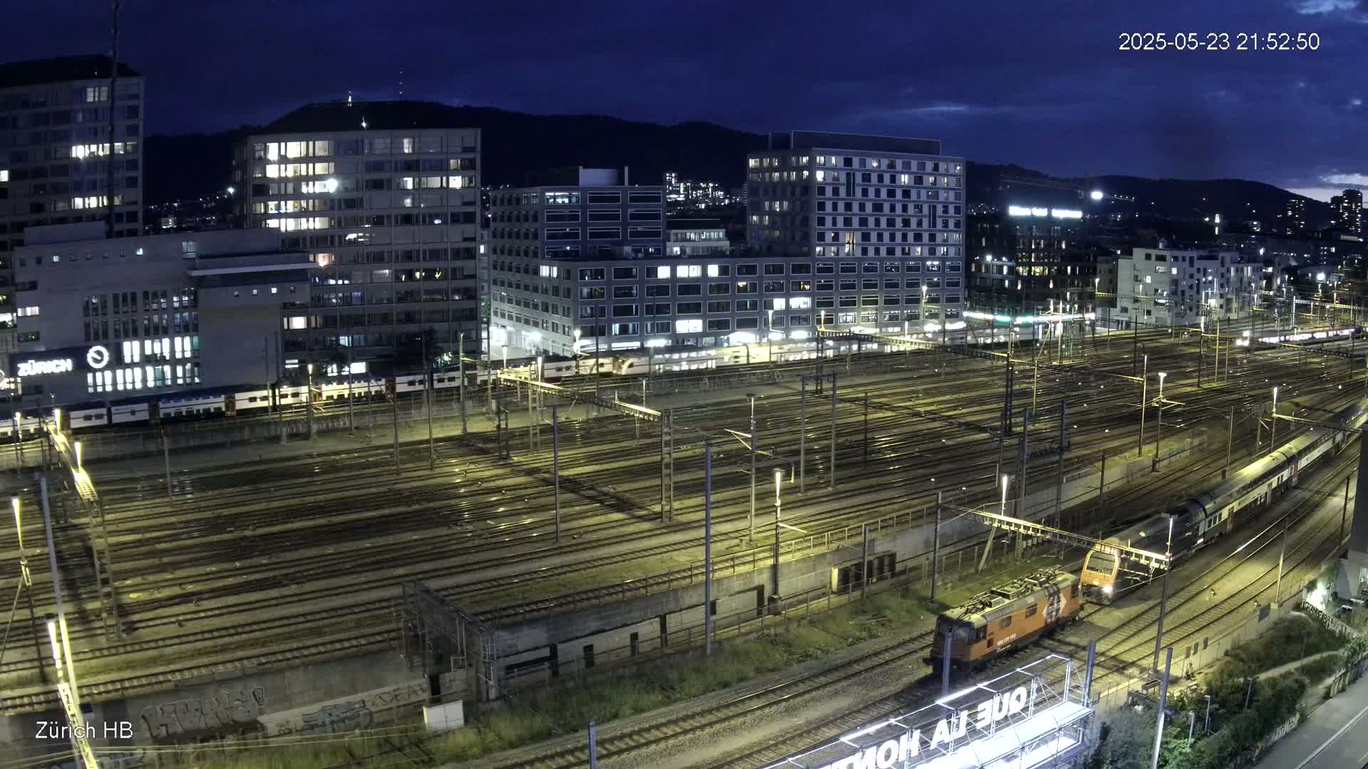 A nighttime aerial view of a large train yard with multiple trains and surrounding city buildings under a dark, clear sky.