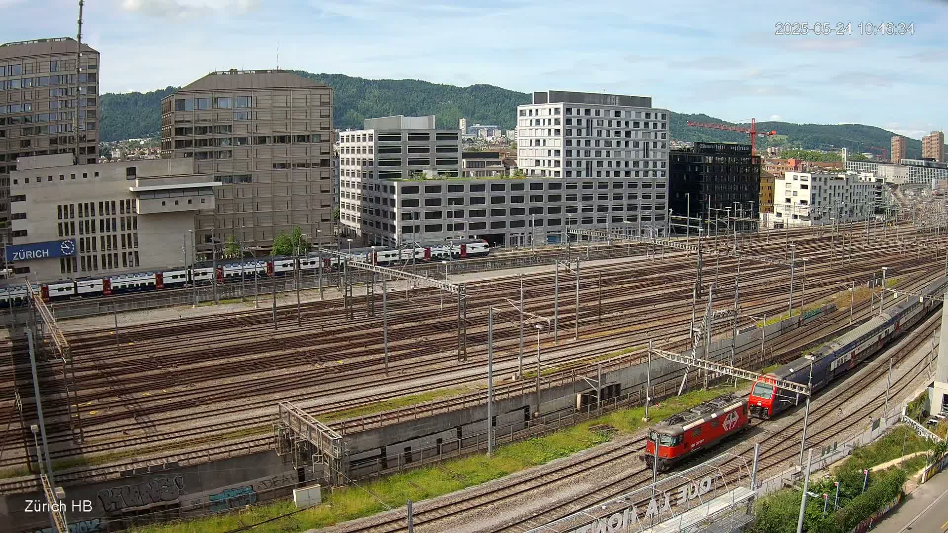 A high-angle view shows a sprawling railway yard with numerous train tracks, several buildings in the background, a partly cloudy sky, and two trains, one red and one blue, visible on the tracks.