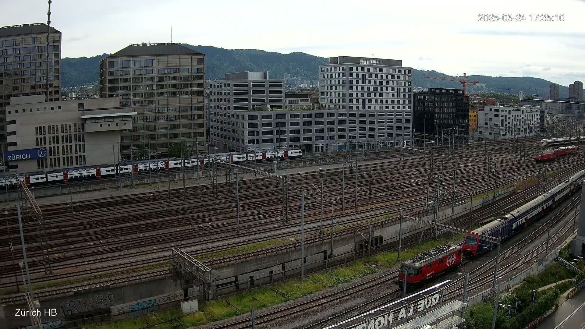 An aerial view shows a sprawling train yard with numerous tracks, several trains, and modern buildings under a partly cloudy sky.