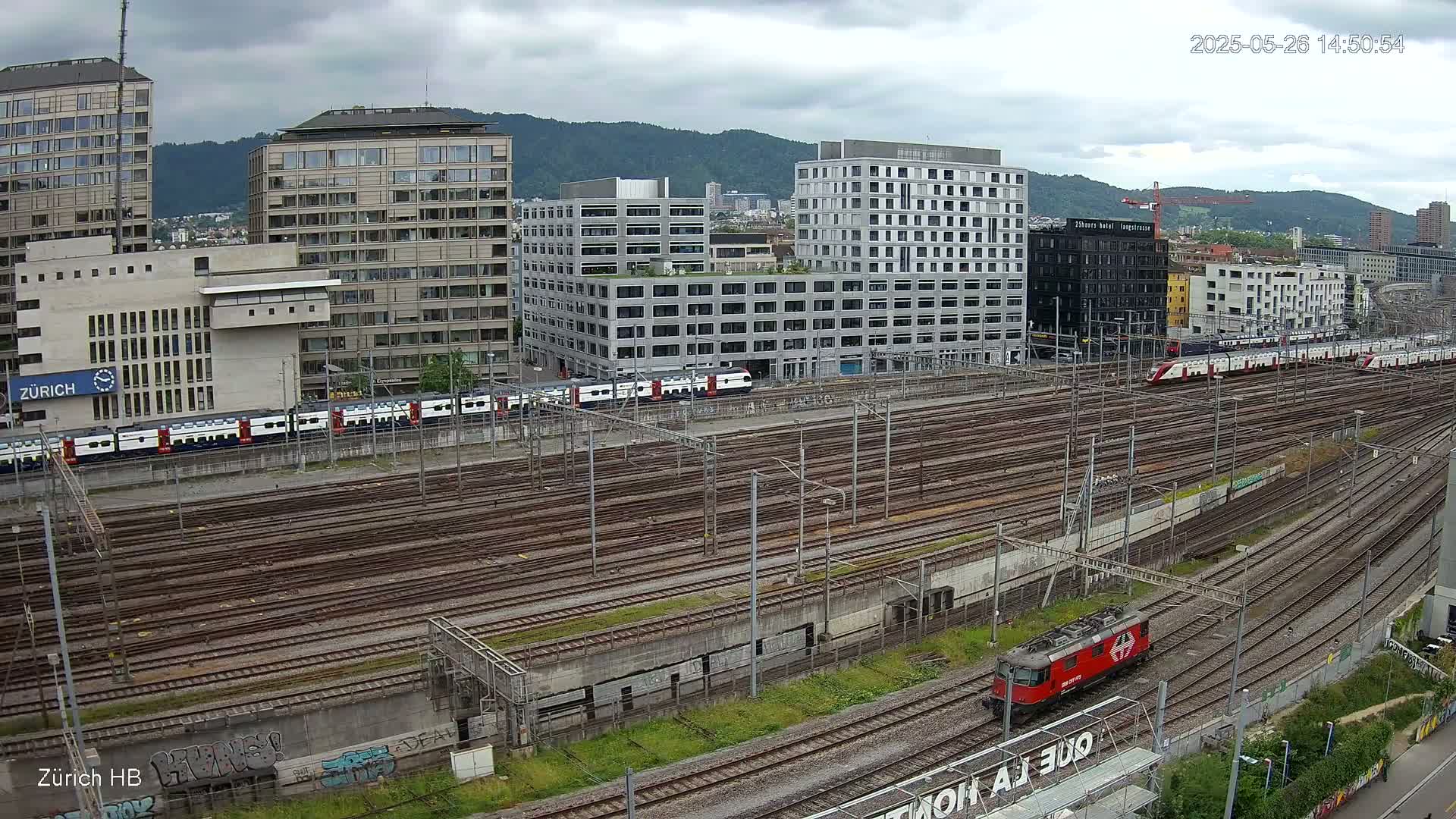 A red locomotive and several passenger trains are visible in a sprawling train yard surrounded by modern buildings under an overcast sky.