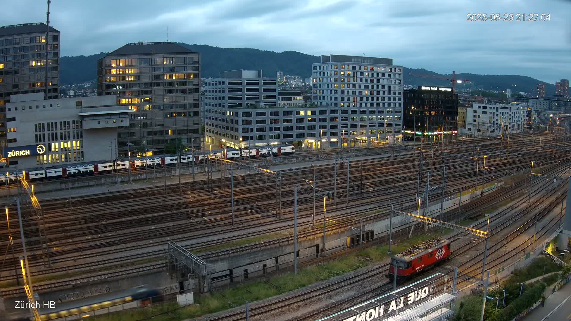A high-angle, twilight view shows a large railway yard with many tracks, several trains, and modern buildings in the background under a cloudy sky.