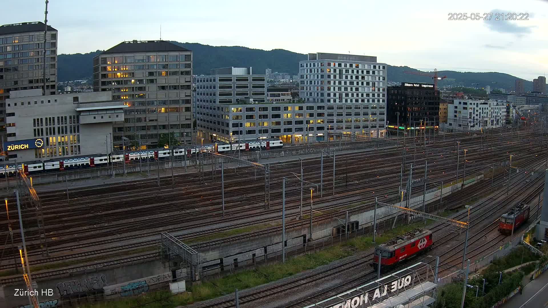 Several buildings are situated near a large network of train tracks at dusk, with two locomotives visible on the tracks.