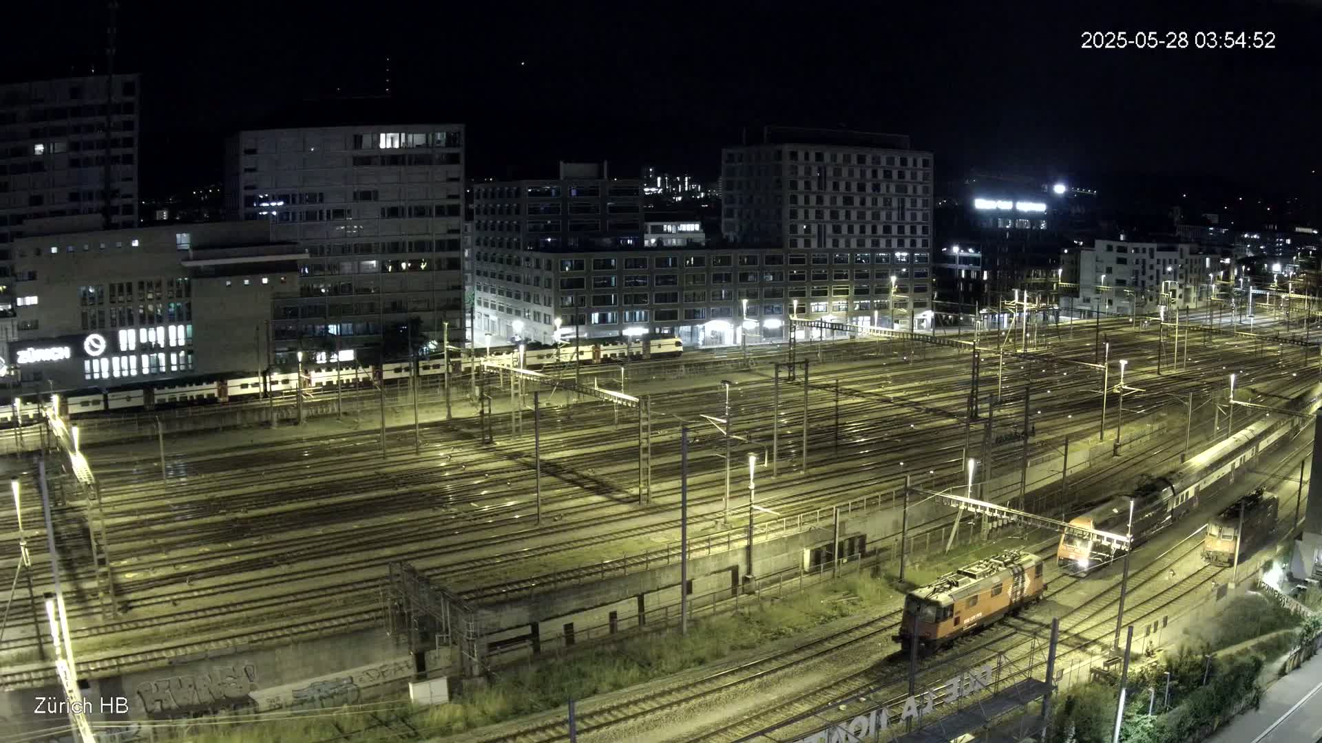 A nighttime, high-angle view shows a sprawling, illuminated train yard with multiple tracks, trains, and surrounding buildings.