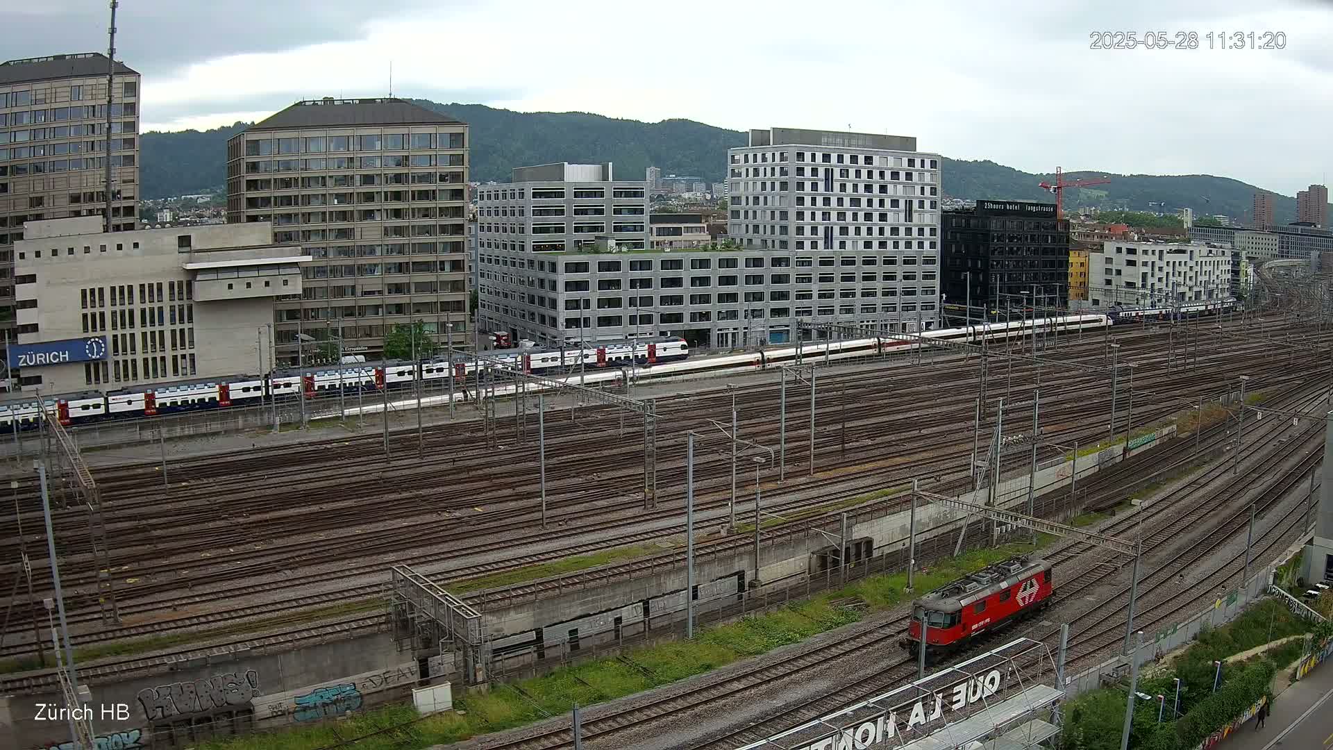 A sprawling railway yard with numerous train tracks, modern buildings, and a long passenger train, under an overcast sky.