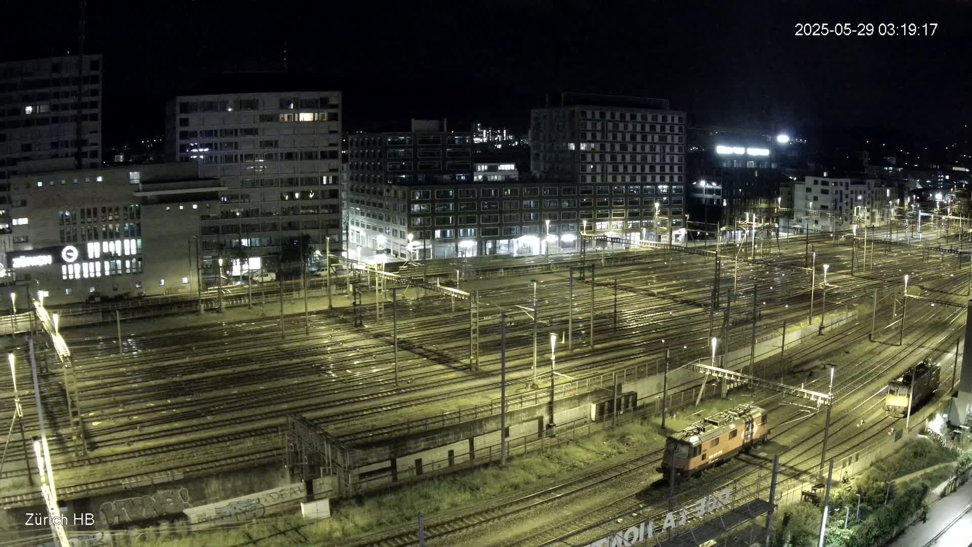 A nighttime aerial view of a large train yard with many tracks and several buildings in the background under clear skies.