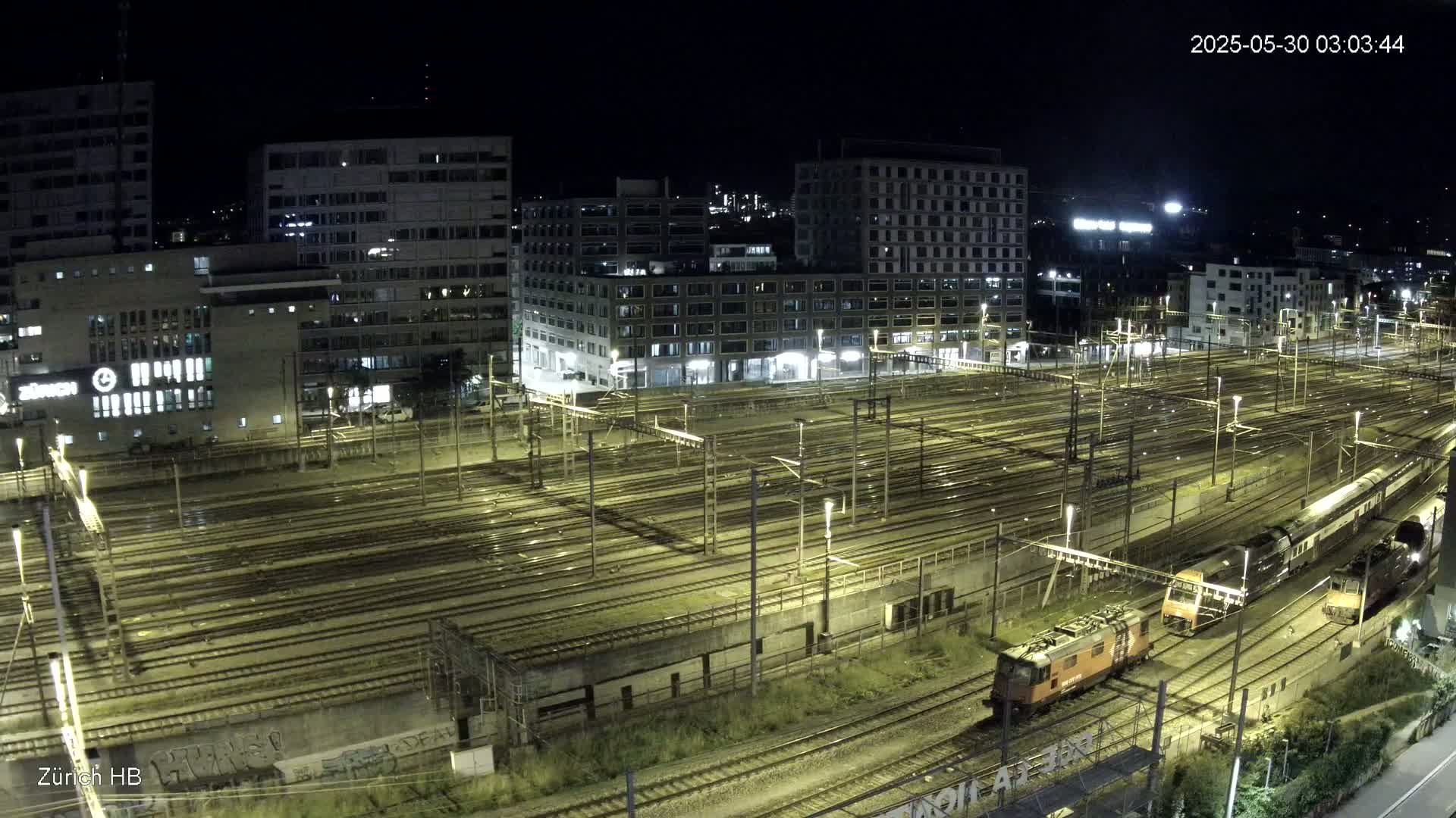 A nighttime, high-angle view of a large train yard with multiple tracks illuminated, surrounded by city buildings.