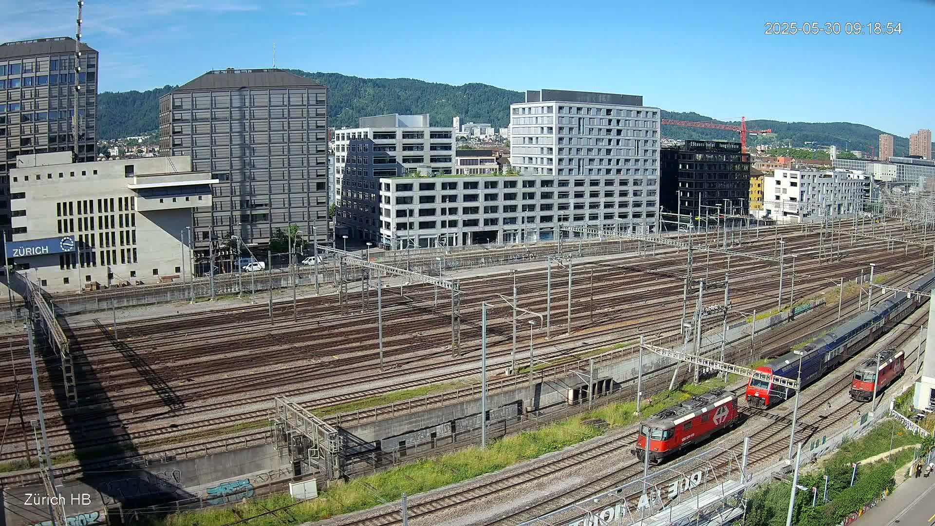 An expansive railway yard with numerous train tracks and several modern buildings in the background under a clear, sunny sky.