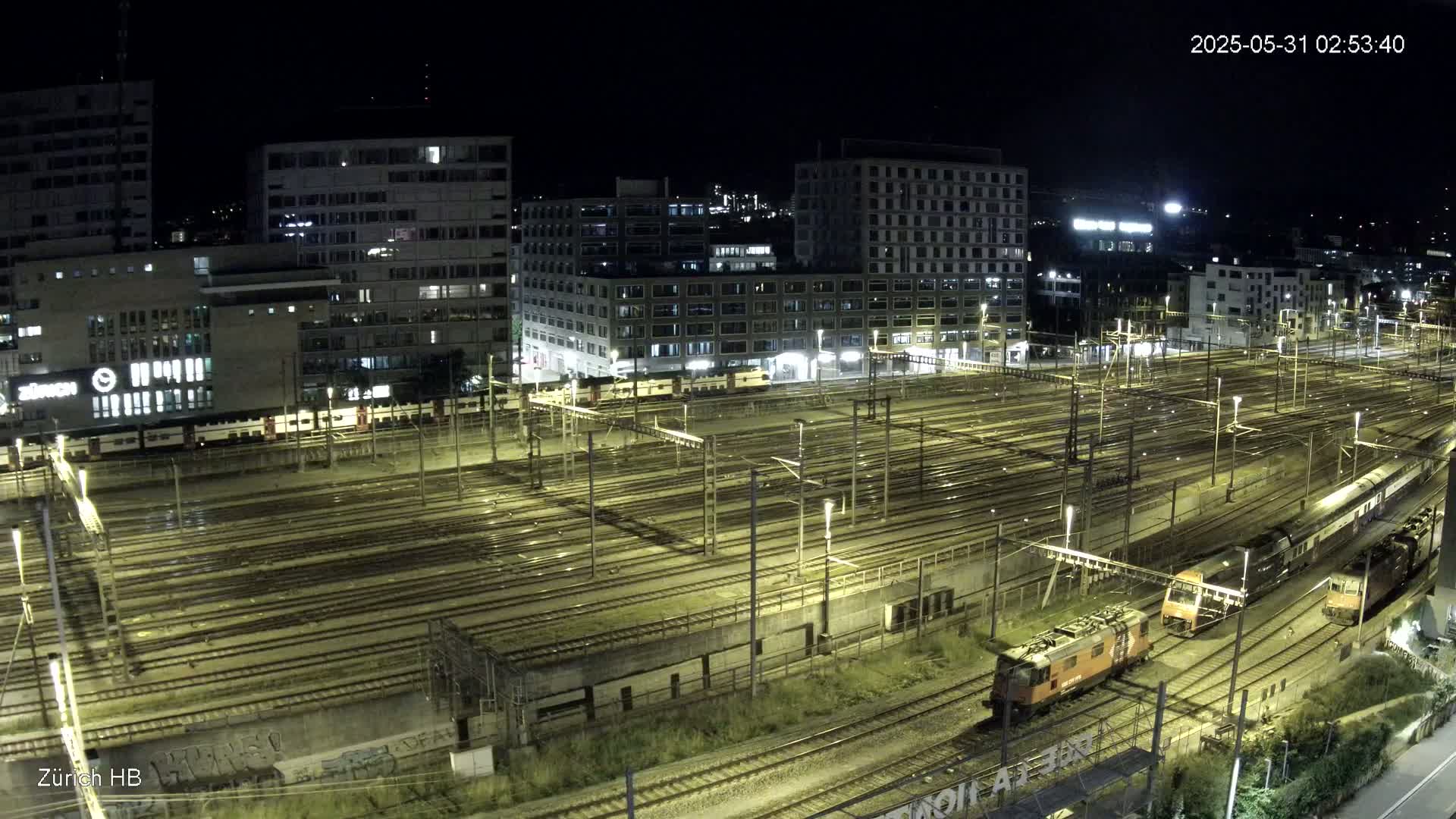 A nighttime, high-angle view of a large railway yard with multiple tracks, trains, and surrounding buildings illuminated under artificial light.