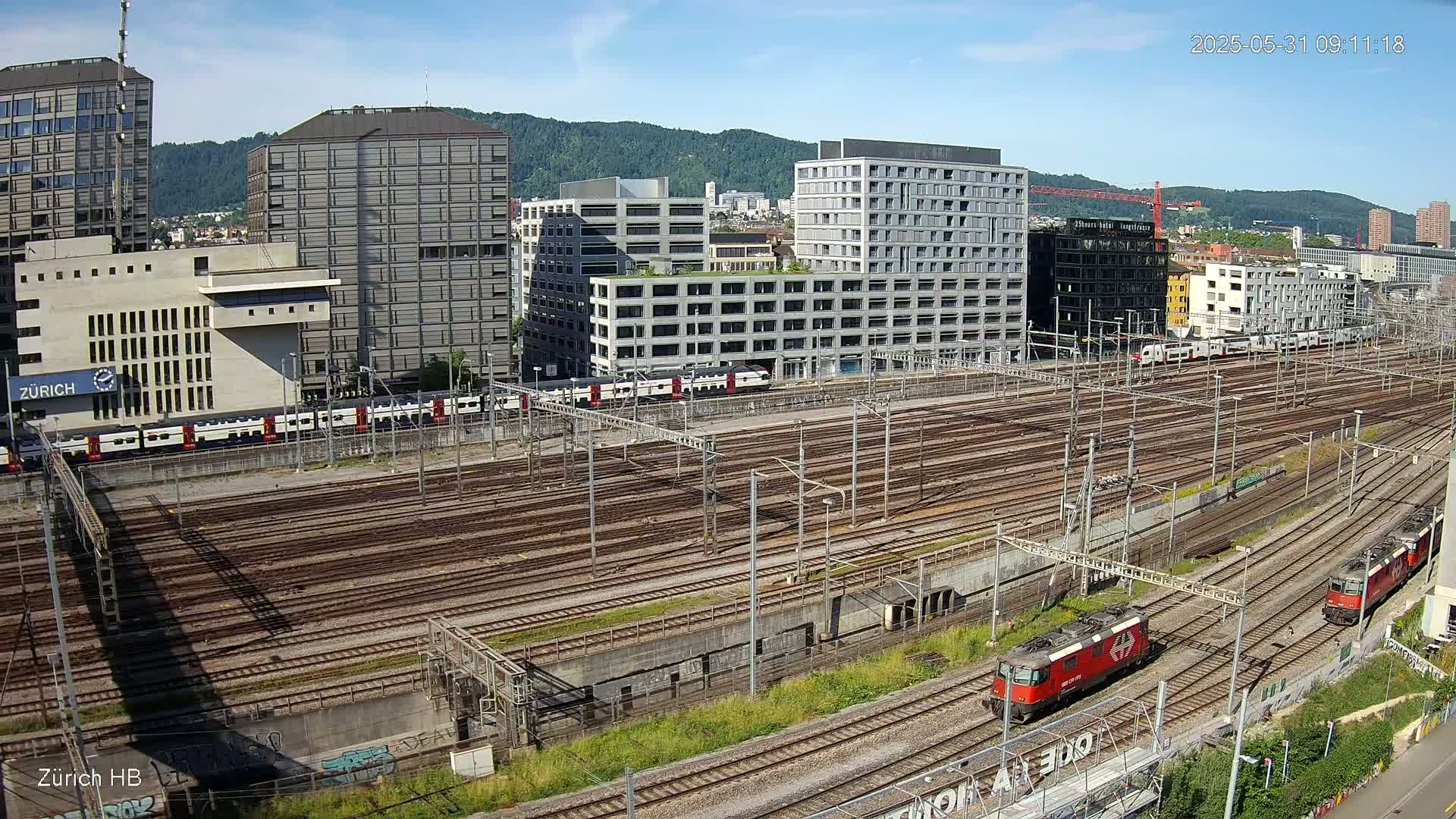 A high-angle view of a sprawling railway yard with numerous train tracks, several trains, and modern buildings in the background under a clear, sunny sky.