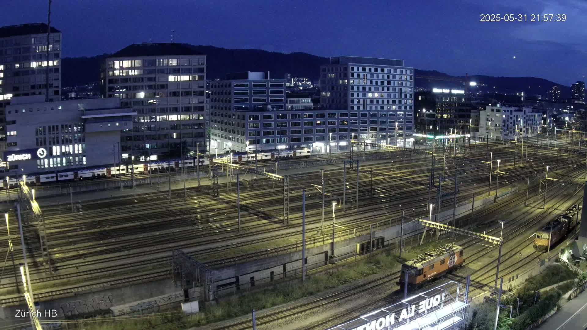 A nighttime, high-angle view shows a sprawling train yard with numerous tracks, several buildings, and two locomotives in the lower right.
