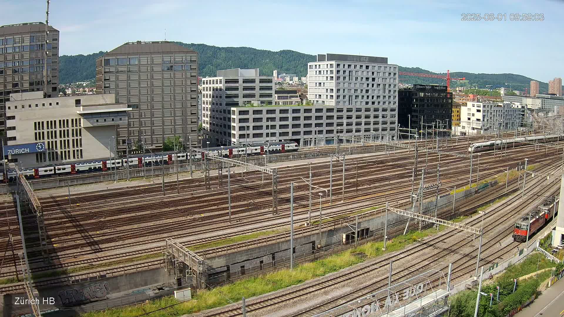 A sunny day view of a large railway yard surrounded by modern buildings and several trains, some in motion.