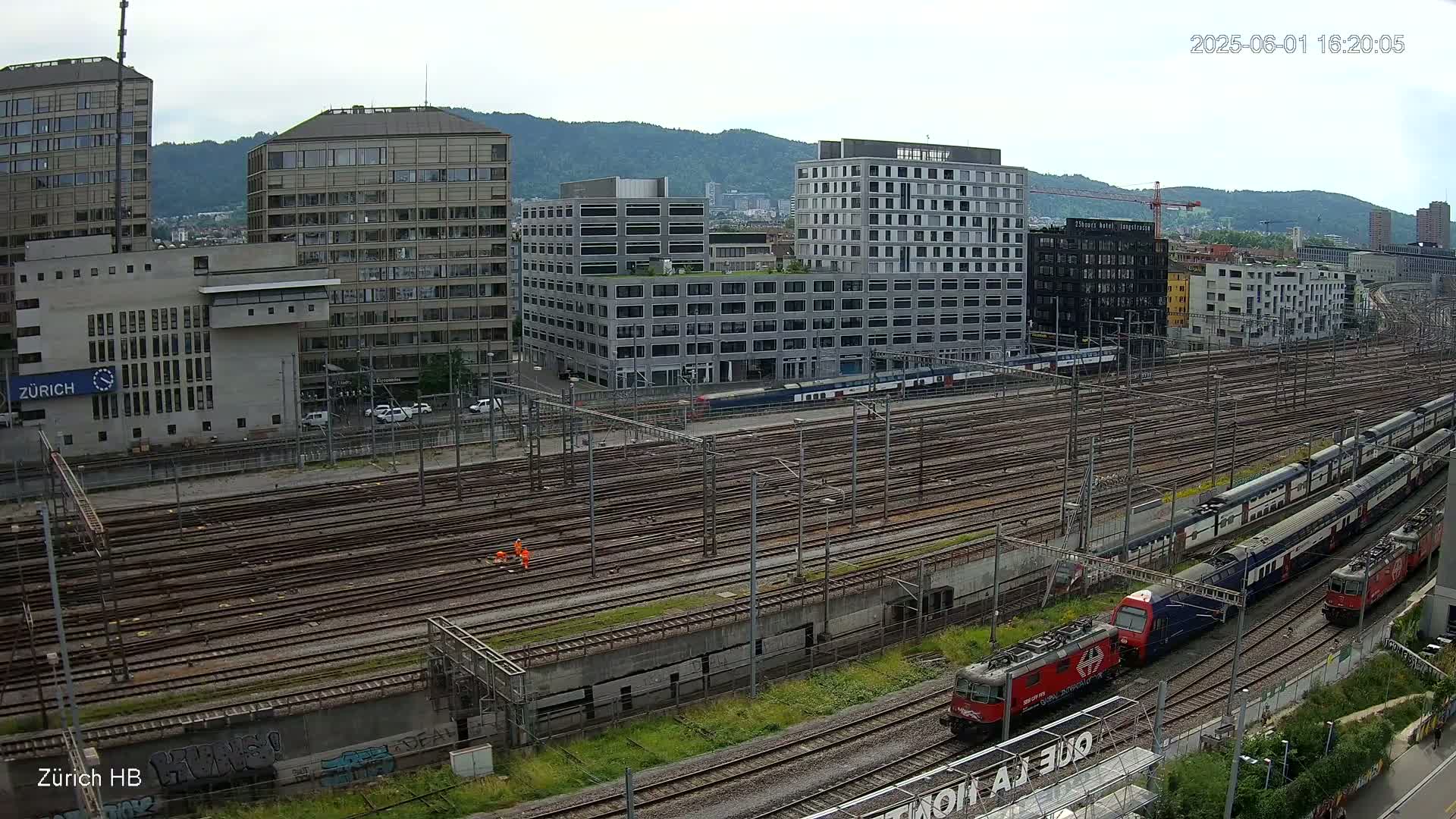 An elevated view of a railway yard with multiple train tracks, several buildings, and mountains in the background on an overcast day.