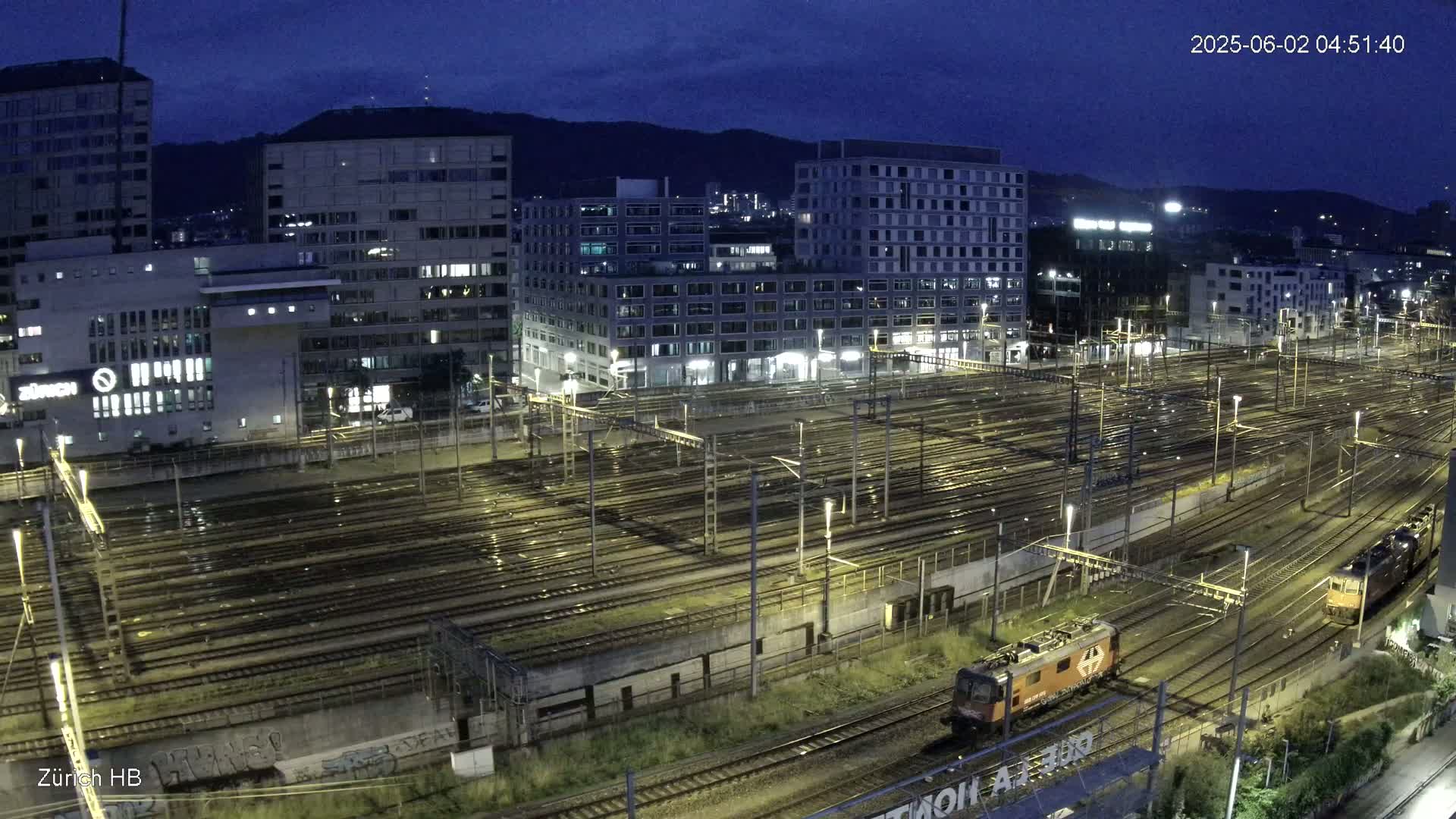 A nighttime, wet view of a large train yard surrounded by city buildings under a dark sky.