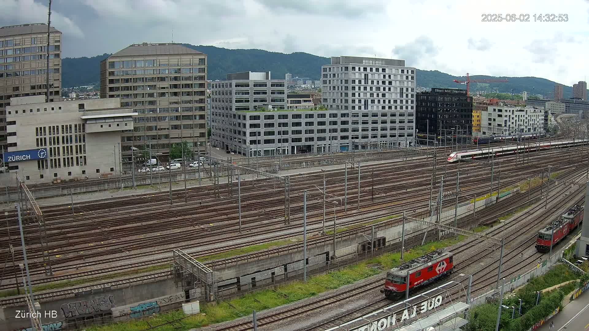 A high-angle view of a railway yard with multiple train tracks, several modern buildings, and two trains, under a partly cloudy sky.