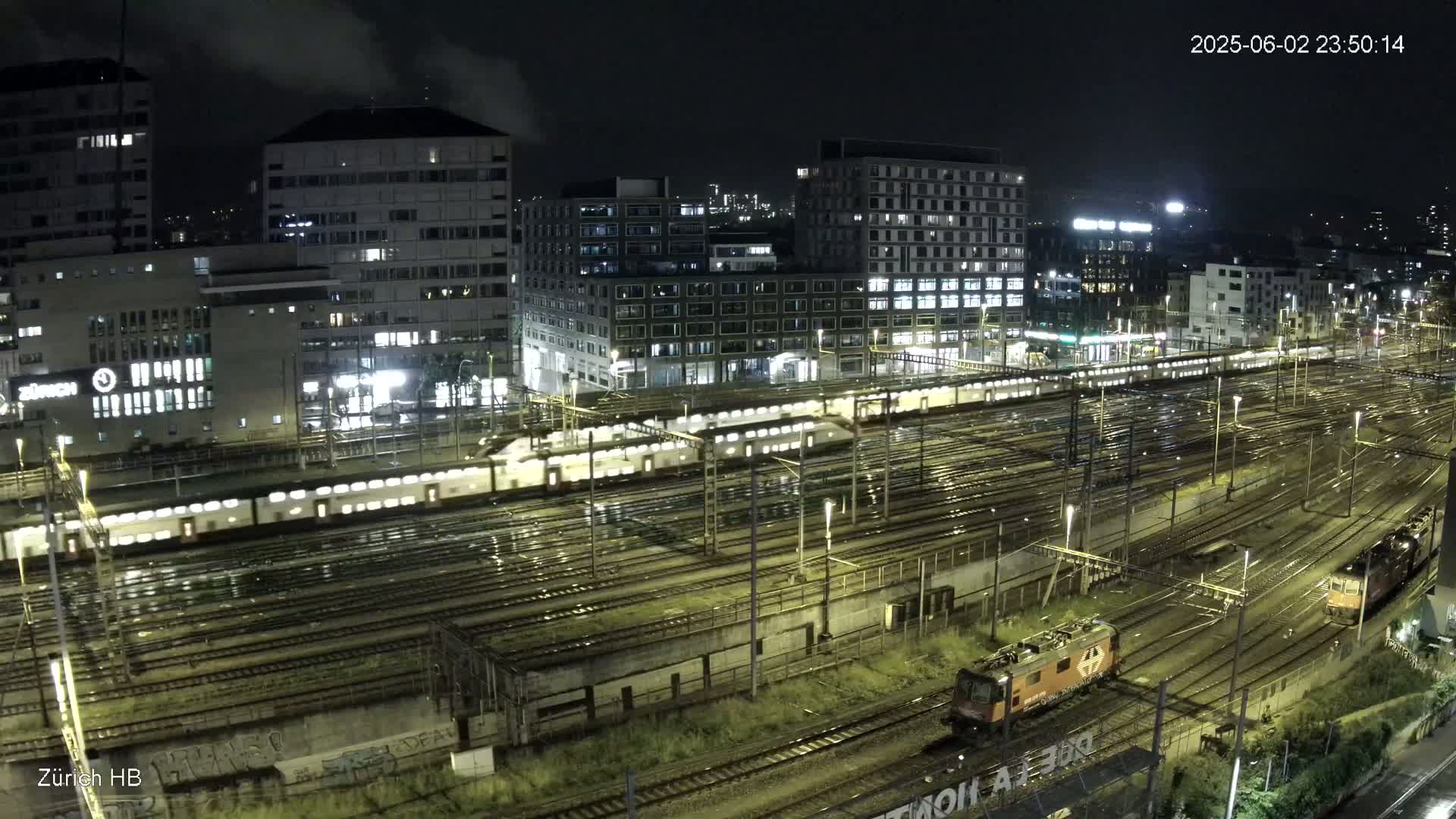 A nighttime view of a rain-slicked train yard with several trains and buildings illuminated in the background.