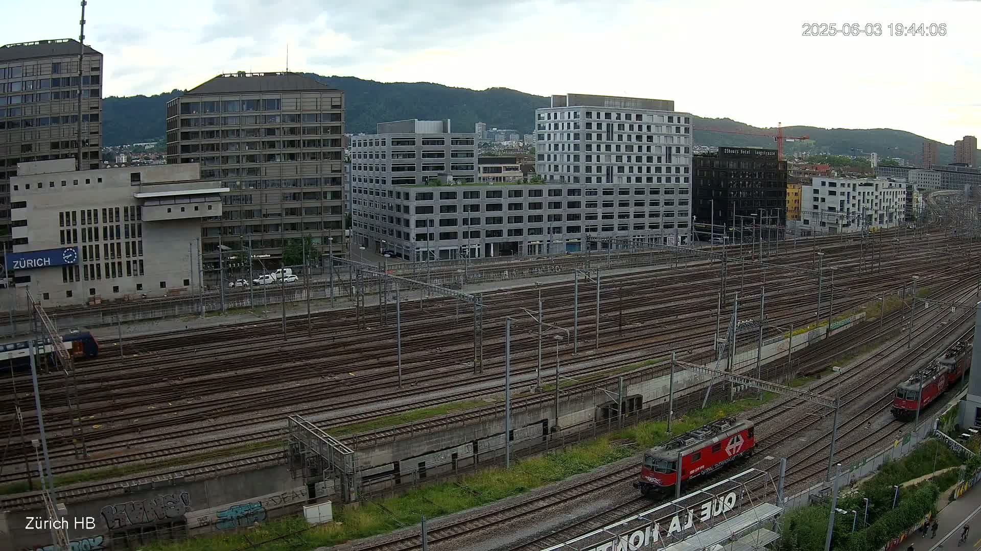 A high-angle view of a railway yard with numerous tracks, surrounded by modern buildings under an overcast sky.
