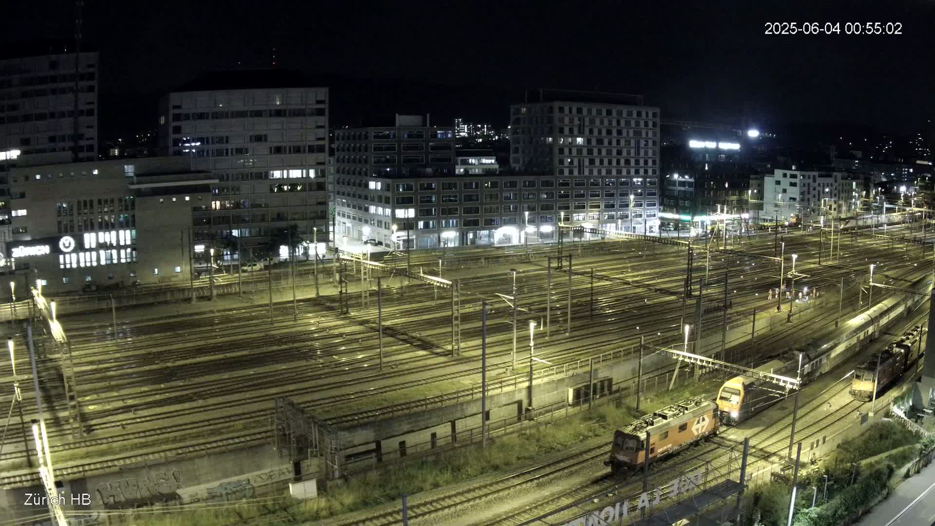 A nighttime high-angle view of a large train yard with numerous tracks and two locomotives, surrounded by city buildings.