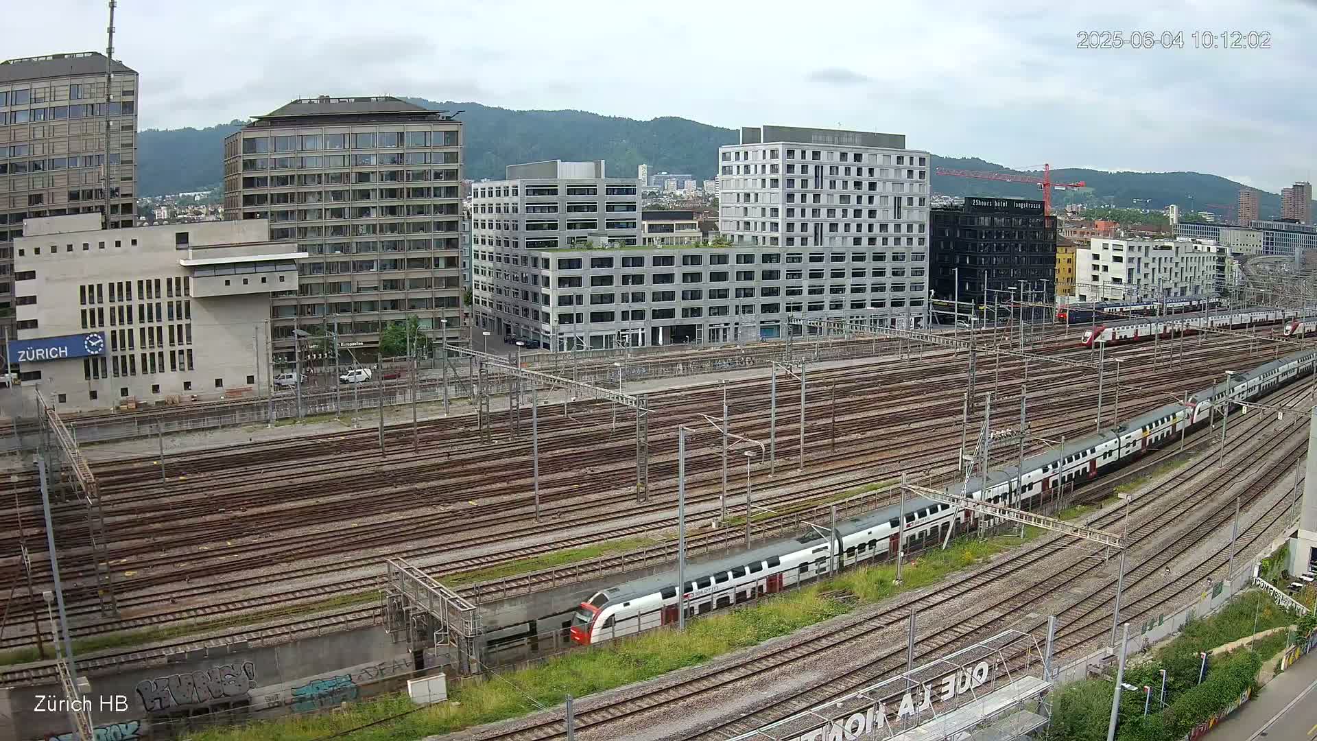 A long passenger train is seen moving through a large railway yard next to several modern buildings under an overcast sky.