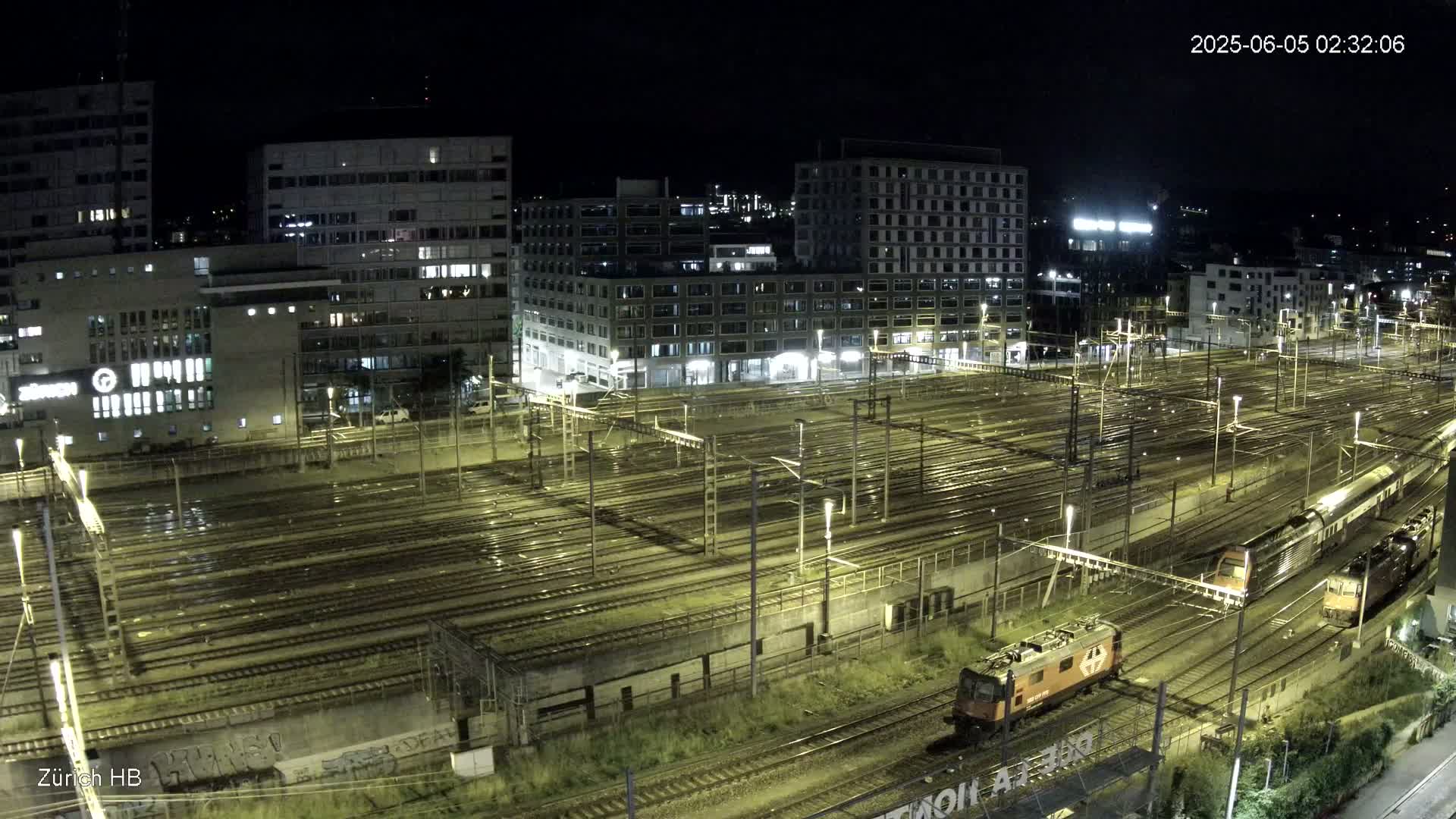 A nighttime, high-angle view of a large train yard with multiple tracks, two locomotives, and surrounding buildings under wet conditions.