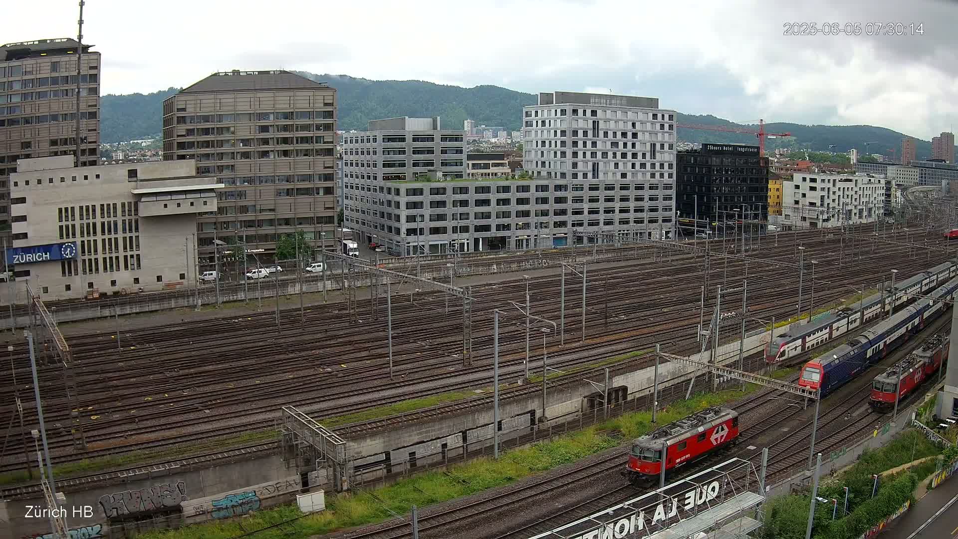A sprawling railway yard with numerous train tracks, surrounded by modern buildings under an overcast sky.