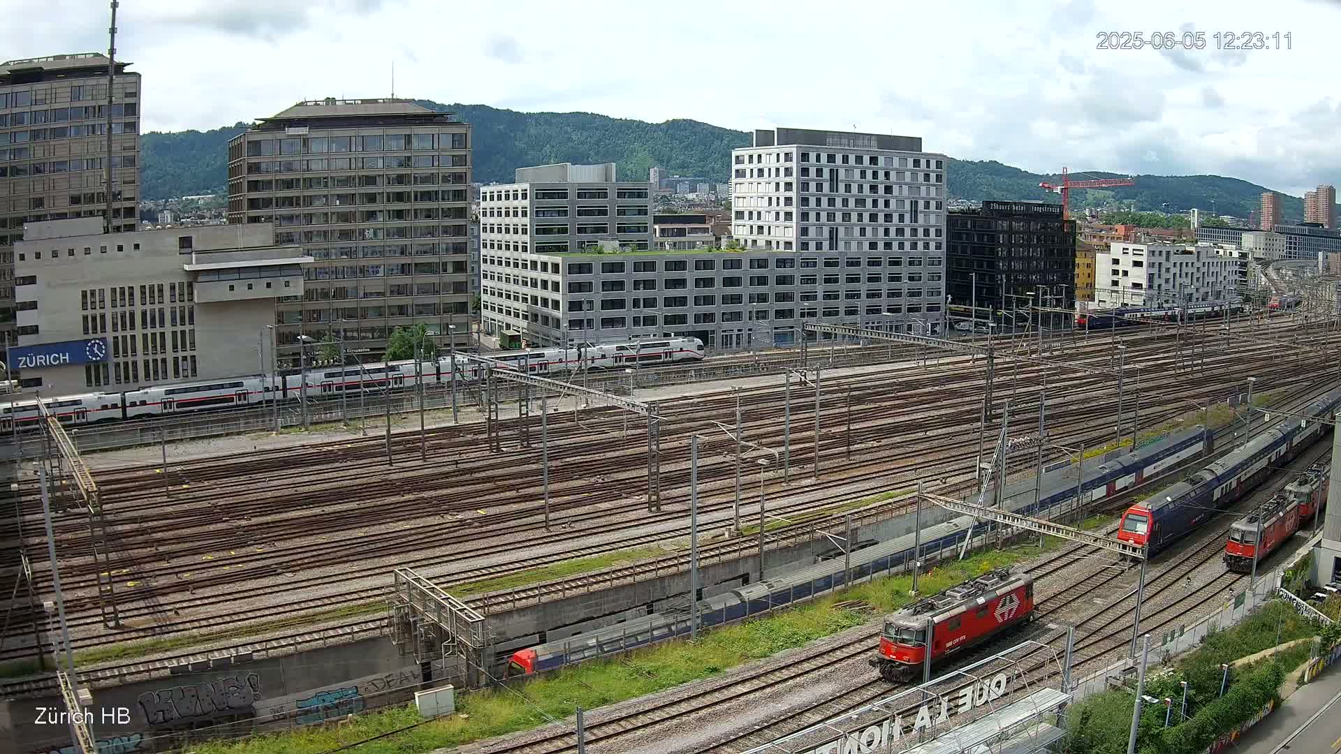 A high-angle view of a sprawling railway yard with numerous train tracks, several trains, and modern buildings in the background under a partly cloudy sky.