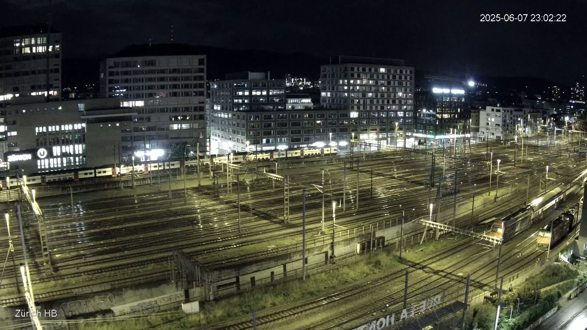A nighttime, wet view of a large train yard surrounded by illuminated buildings.