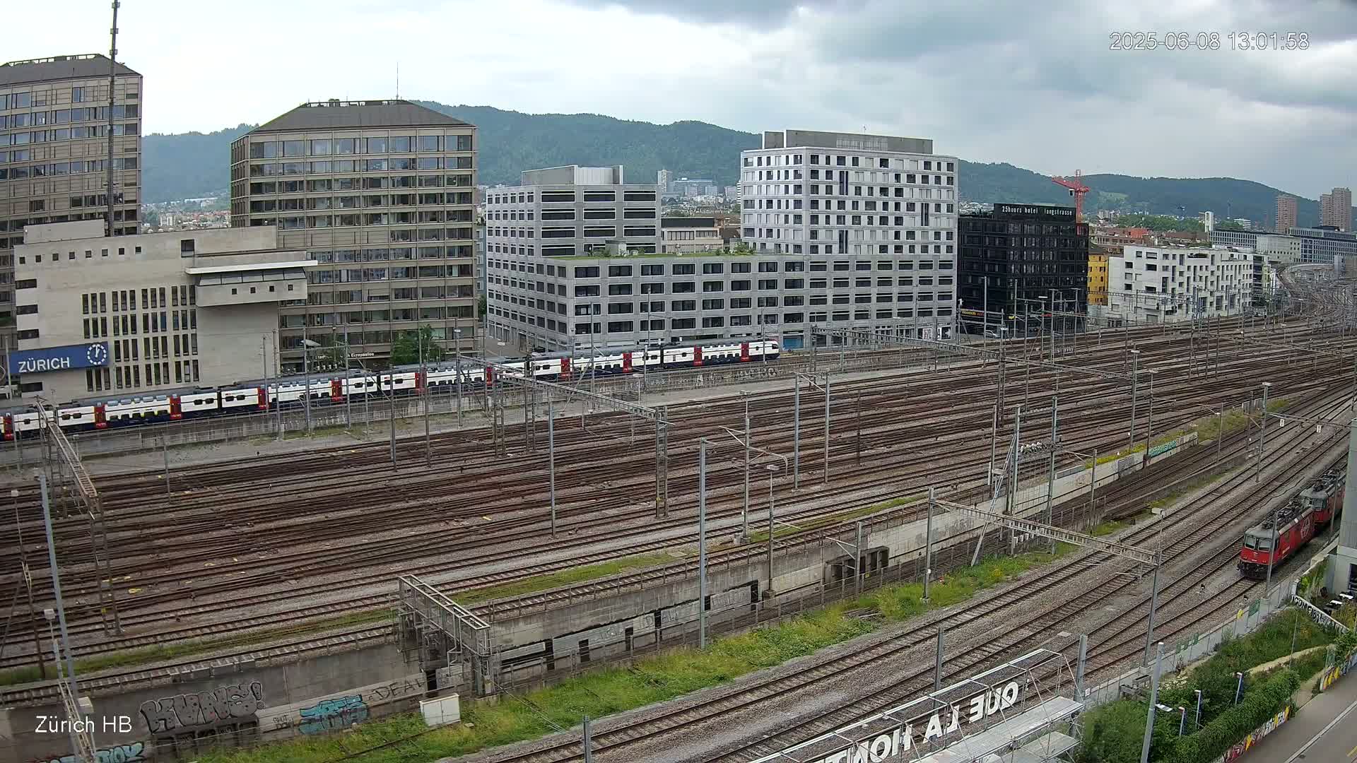 A wide shot of a railway yard with numerous train tracks, surrounded by modern and older buildings under a cloudy sky.