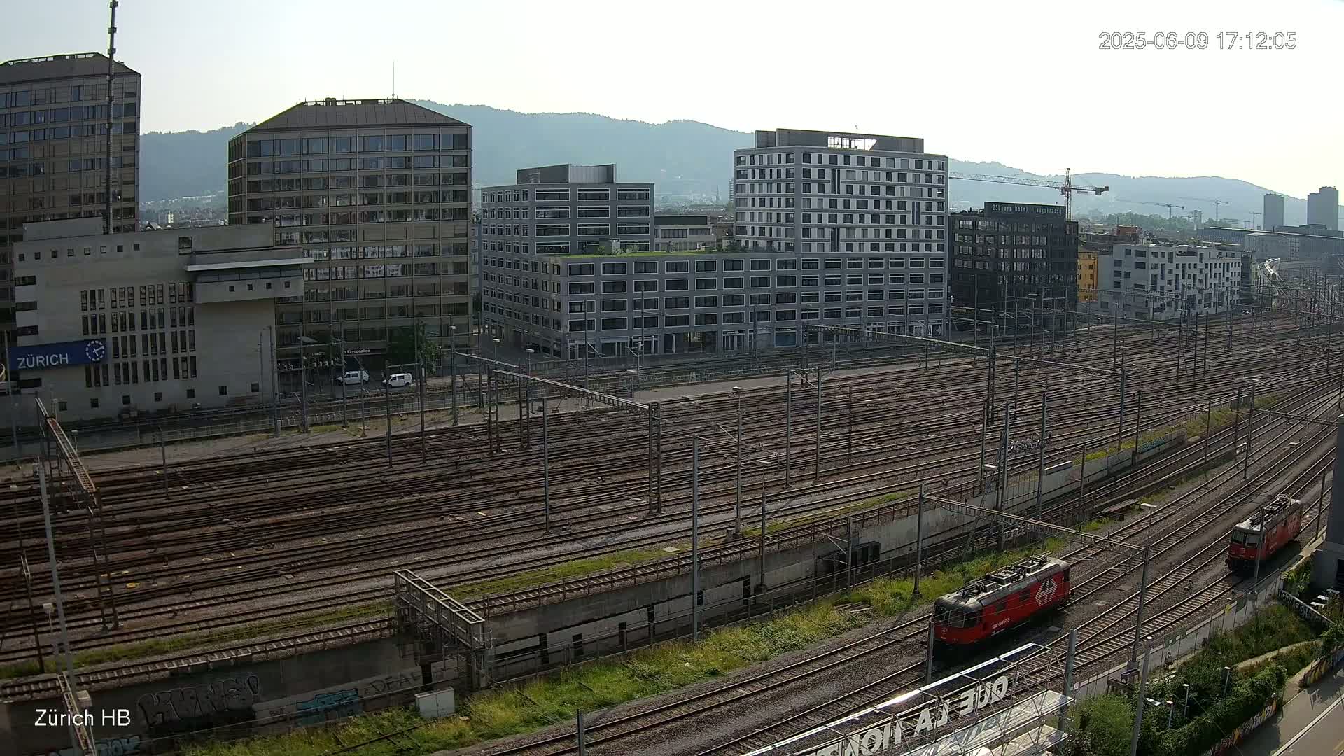 An aerial view shows a large railway yard with many tracks, two locomotives, and several multi-story buildings on a sunny day.