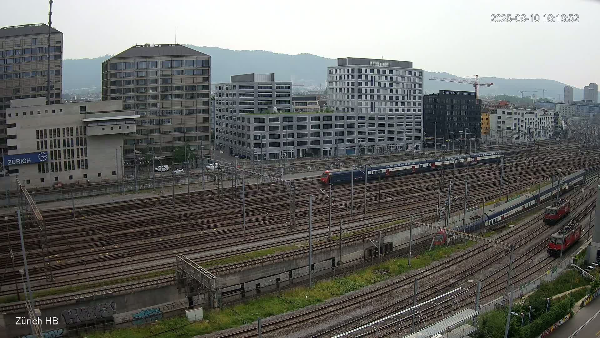 A high-angle view shows a sprawling railway yard with numerous train tracks, several trains, and modern buildings on a cloudy day.