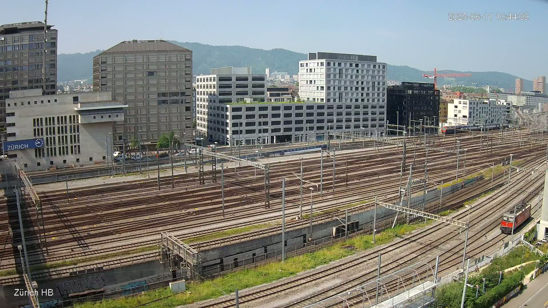 A sprawling railway yard with numerous tracks and several modern buildings in the background on a clear, sunny day.