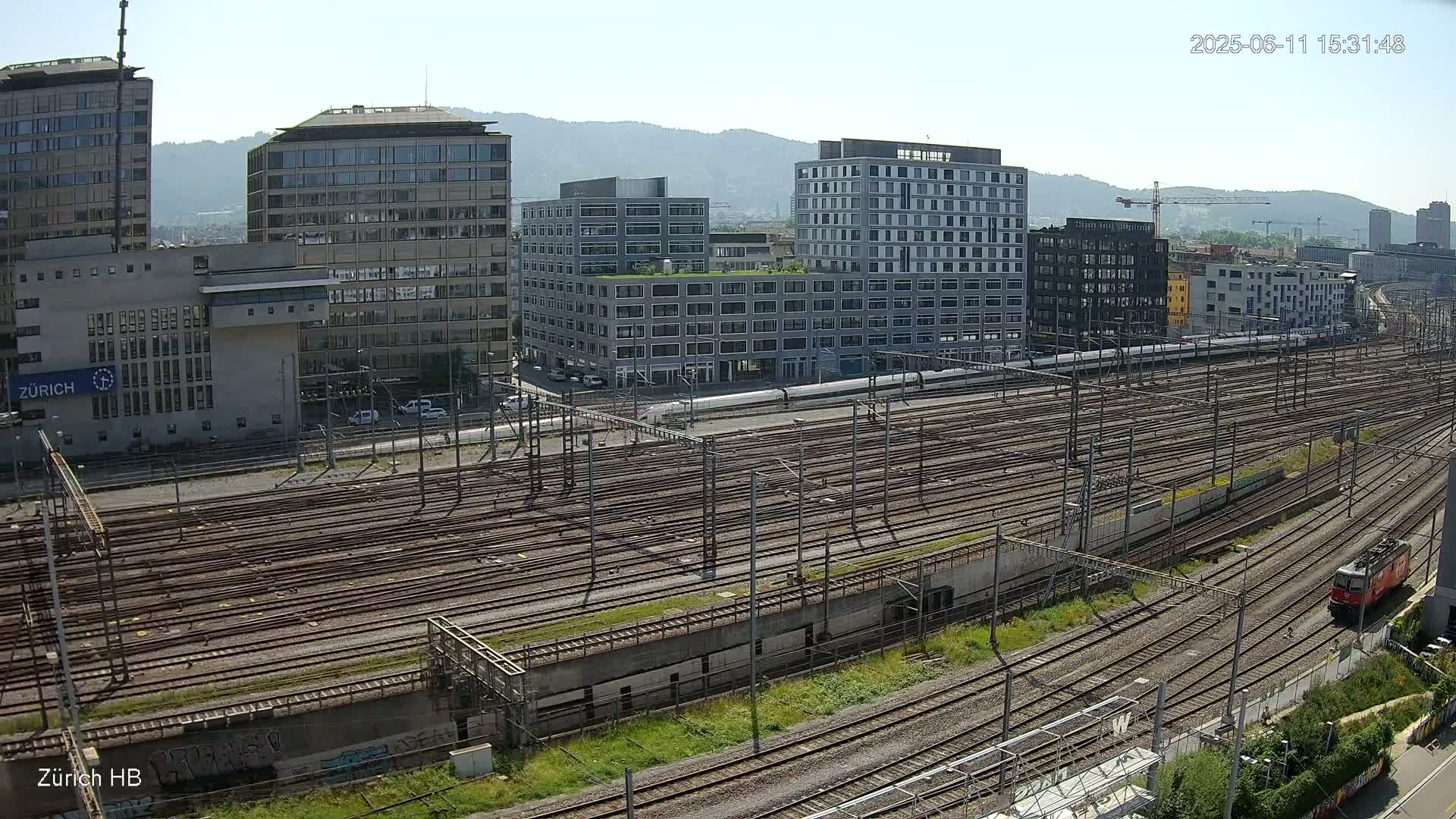 A high-angle view on a sunny day shows a large railway yard with numerous tracks, surrounded by modern and older buildings, including high-rises, and a train passing through.