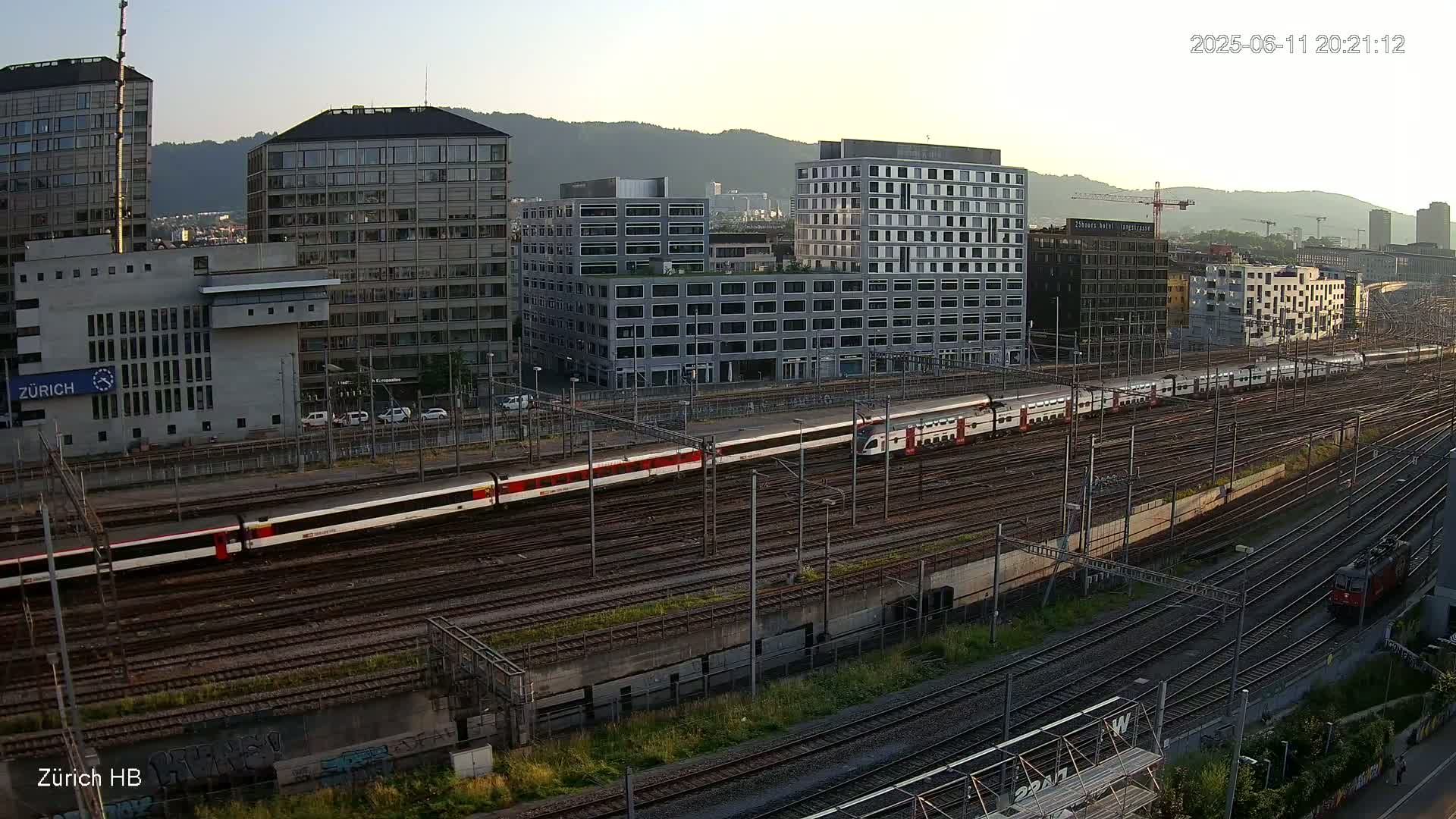 A long passenger train travels across a railway yard next to several modern buildings under a clear, twilight sky.