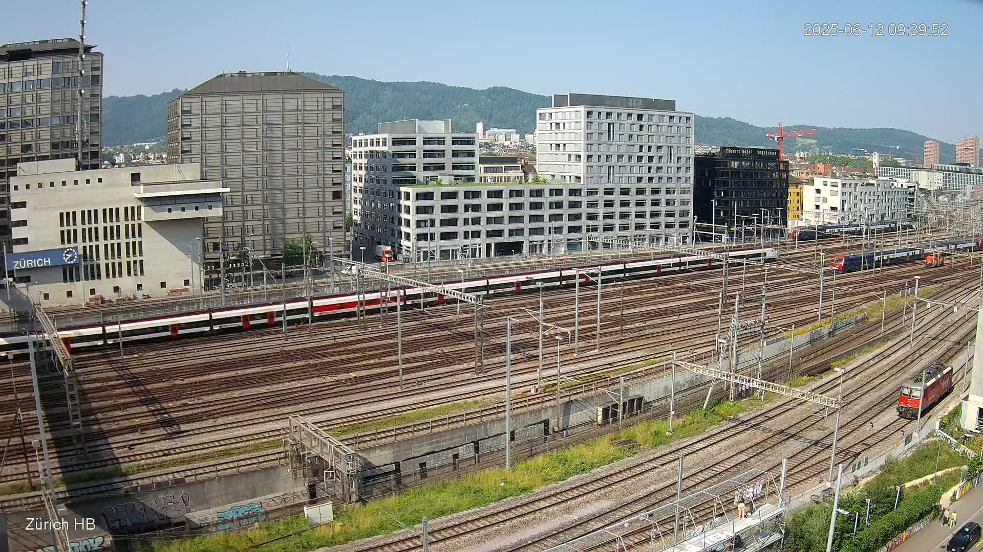 A sunny day view of a large train yard with multiple trains and surrounding city buildings.
