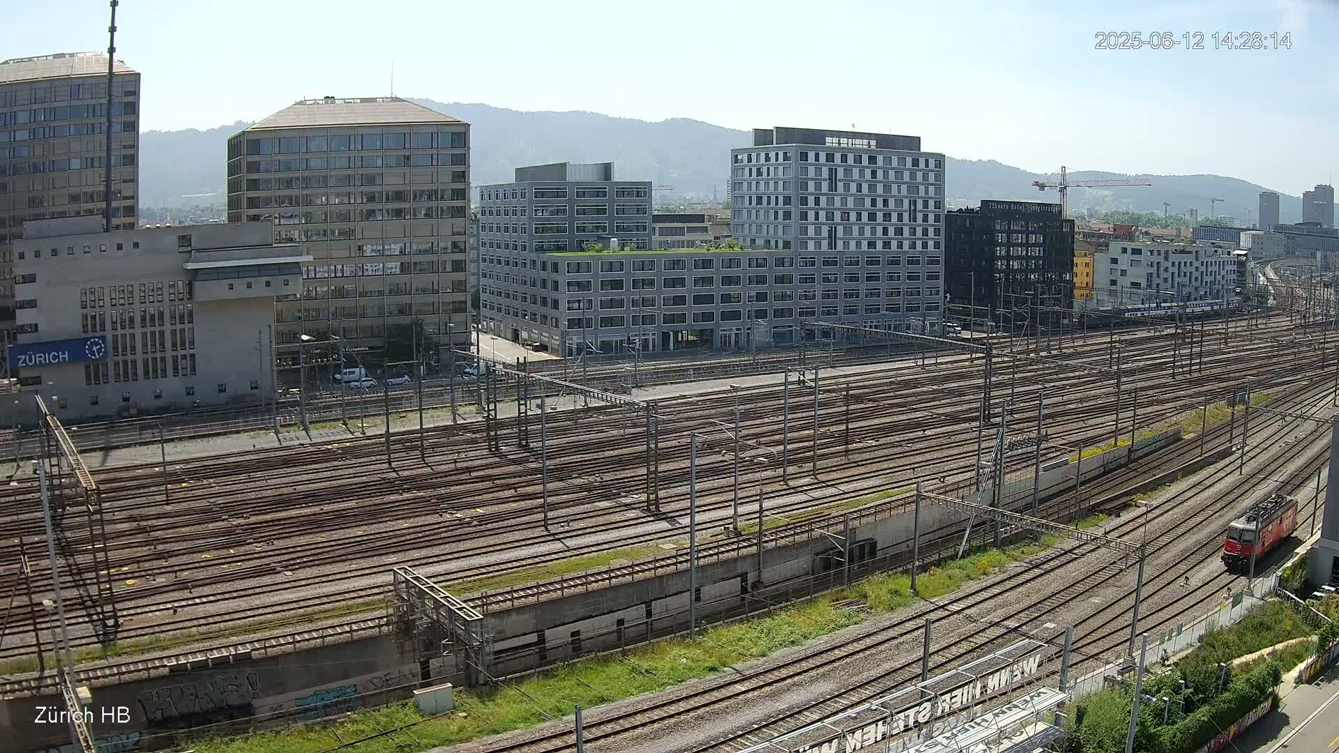 A high-angle view on a sunny day shows a large railway yard with many tracks surrounded by modern buildings and a train.