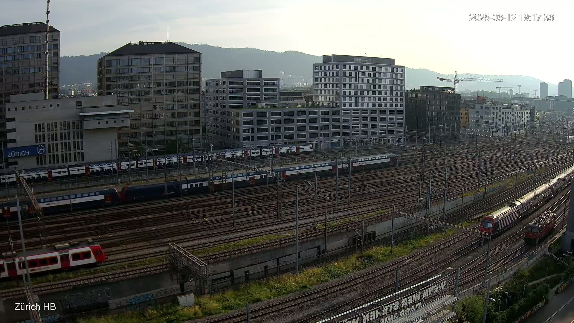 Several trains are parked and moving on a large railway network surrounded by modern buildings under a hazy sky.