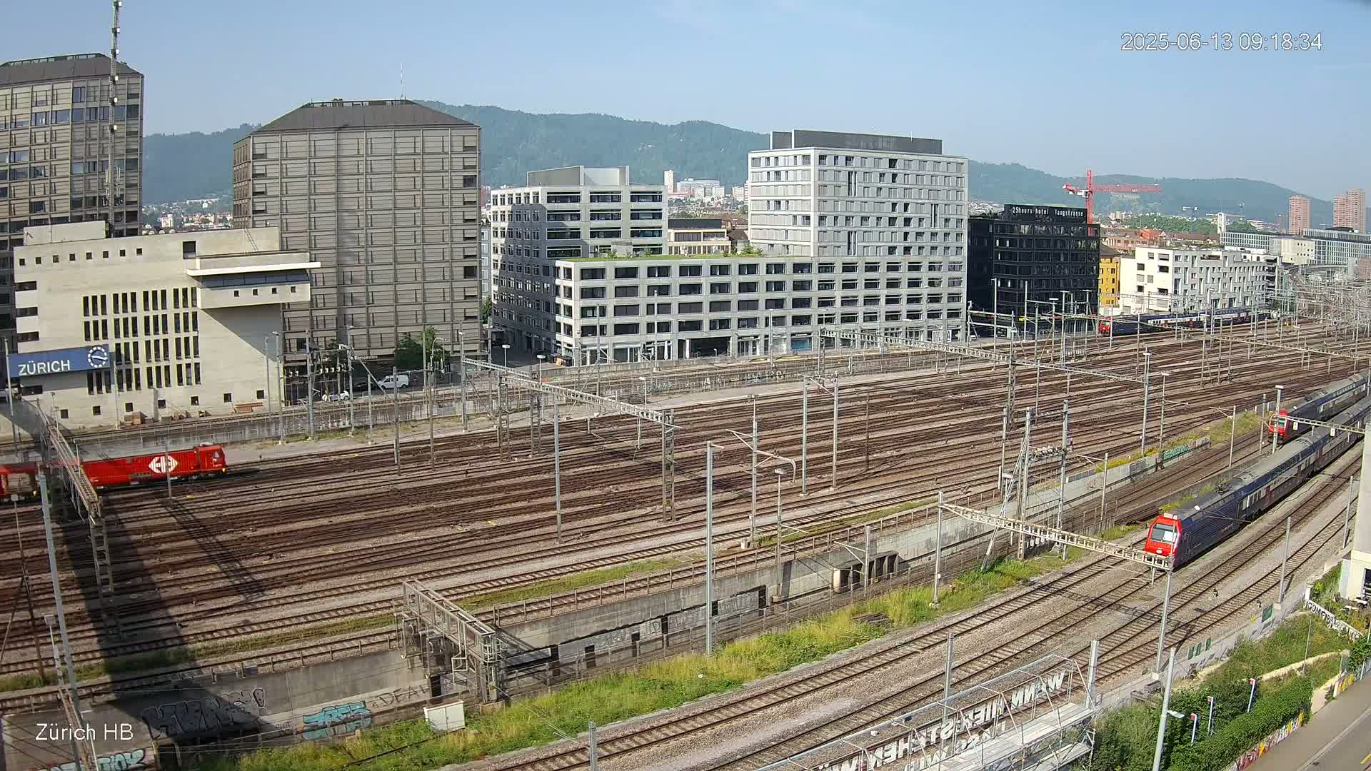 A high-angle view of a large railway yard with numerous tracks, surrounded by modern buildings under a clear, sunny sky.