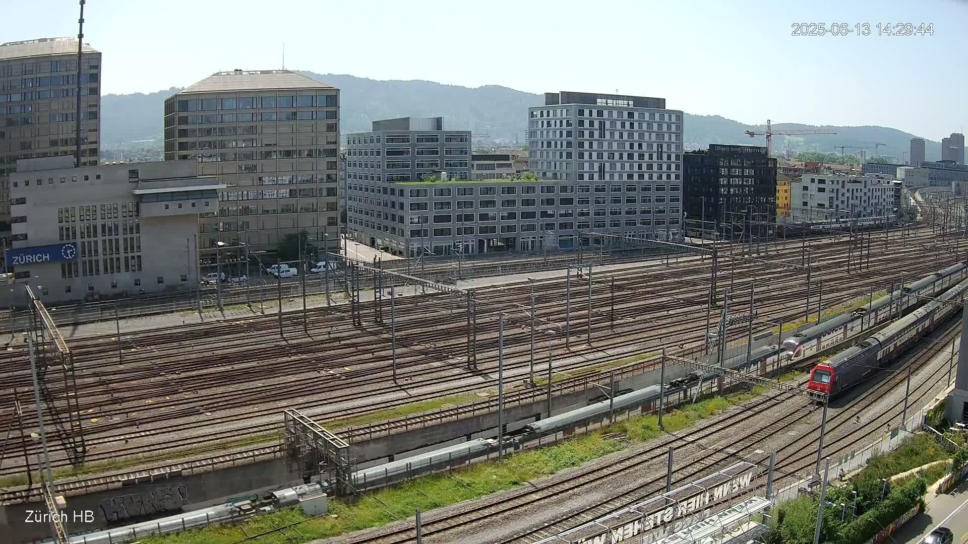 A high-angle view of a train station on a sunny day shows numerous train tracks extending into the distance, with several trains visible and buildings in the background.