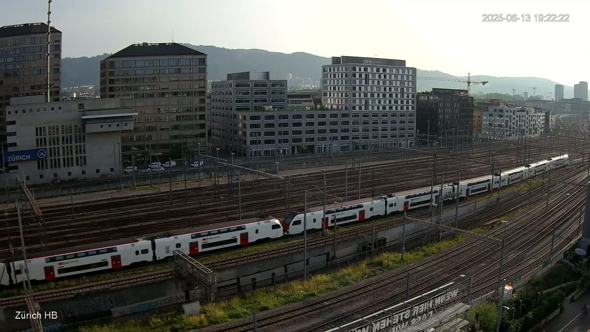 A long white and red train travels along a network of train tracks in front of several multi-story buildings under a hazy sky.