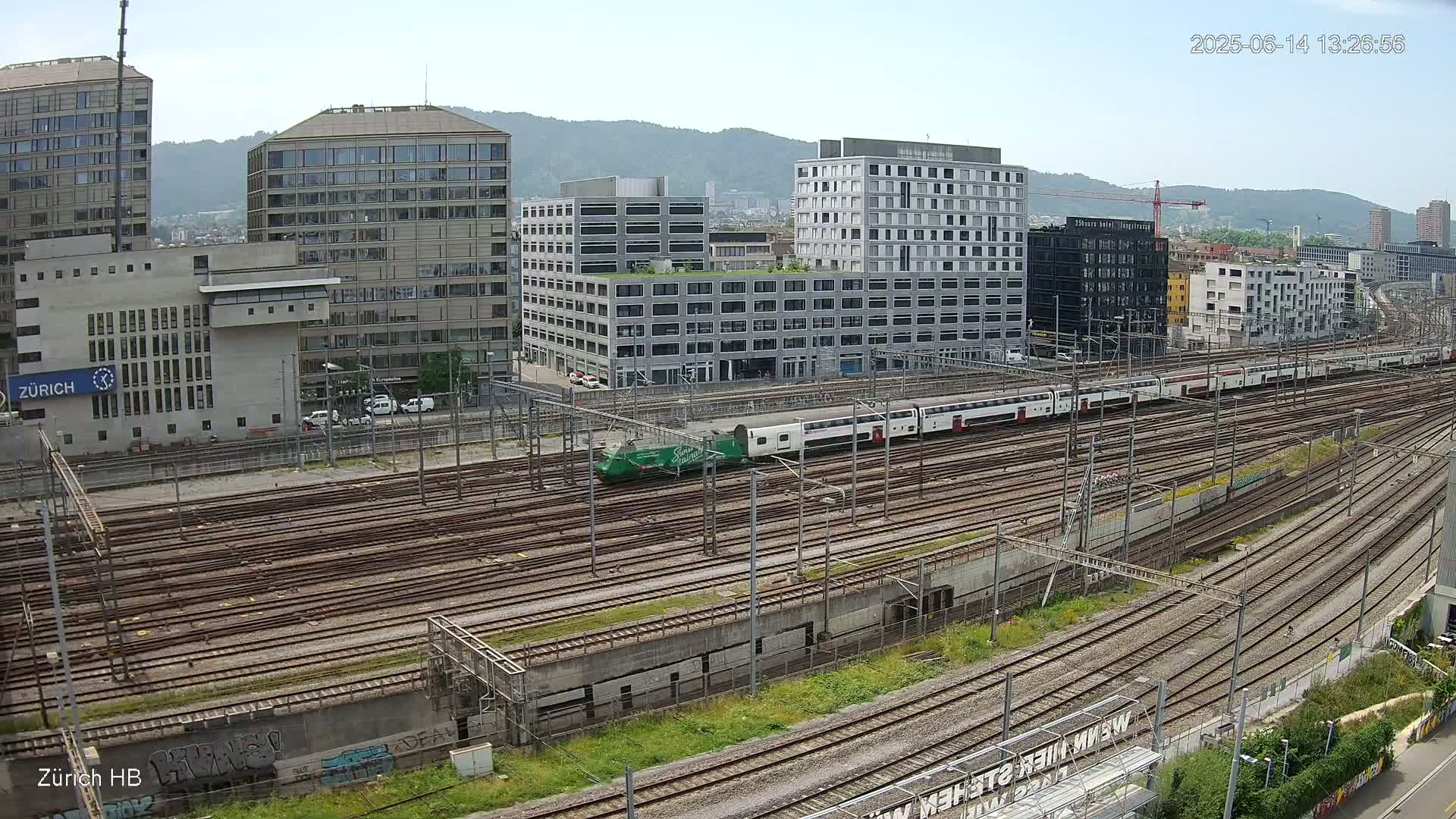 A long expanse of train tracks stretches out before several modern buildings under a bright, sunny sky.