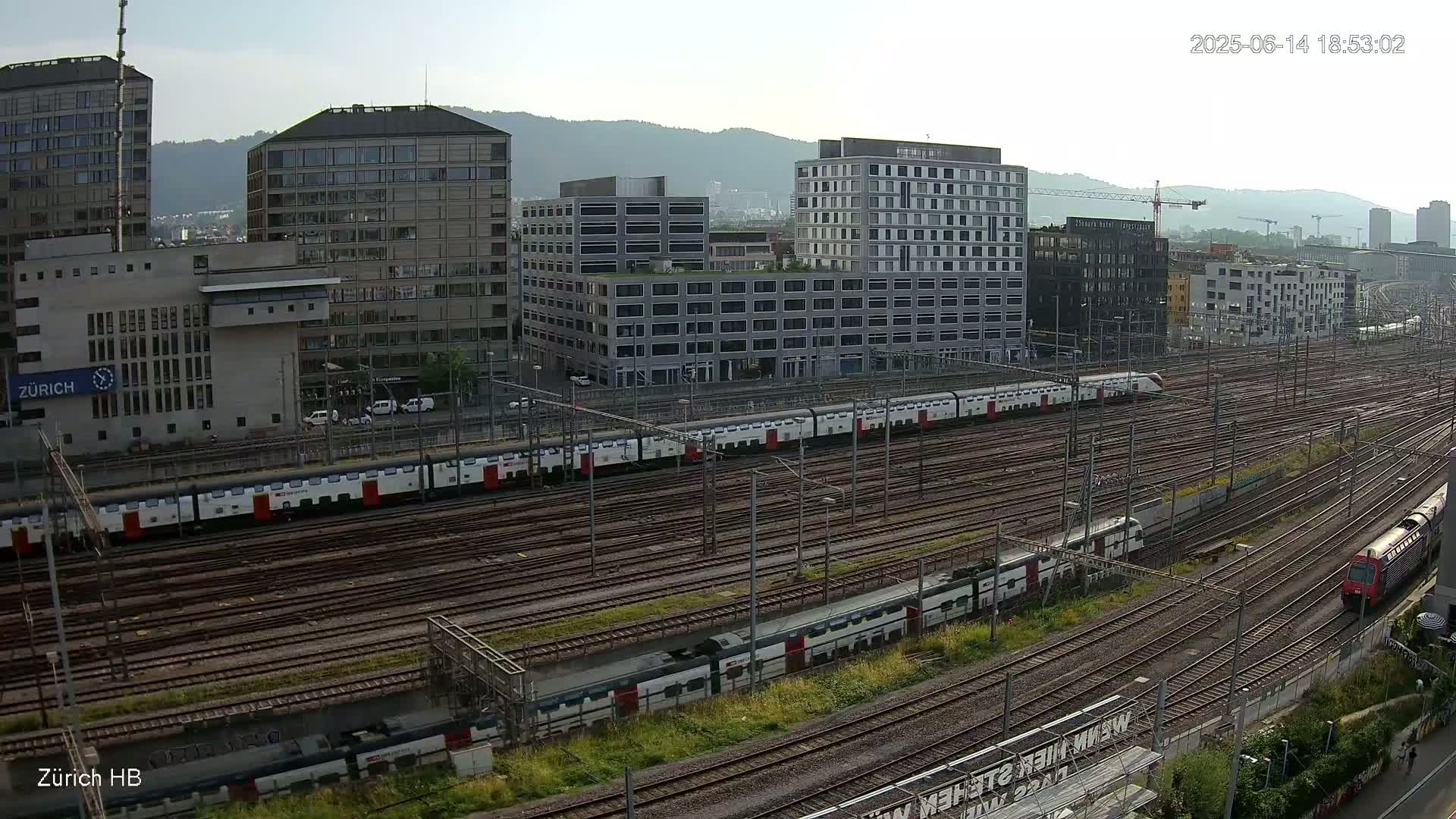 A long passenger train sits on a railway network next to several modern buildings under a clear, sunny sky.