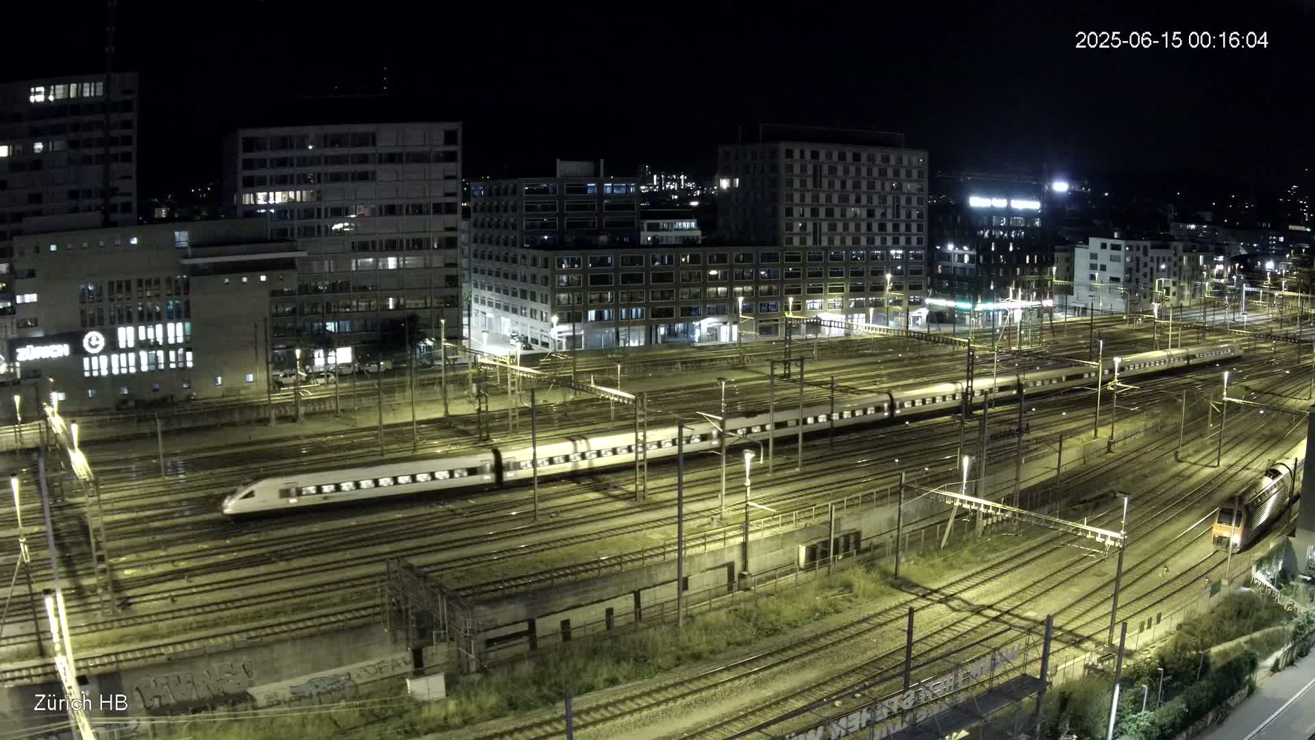 A long passenger train sits on a railway network next to several modern buildings under a clear, sunny sky.